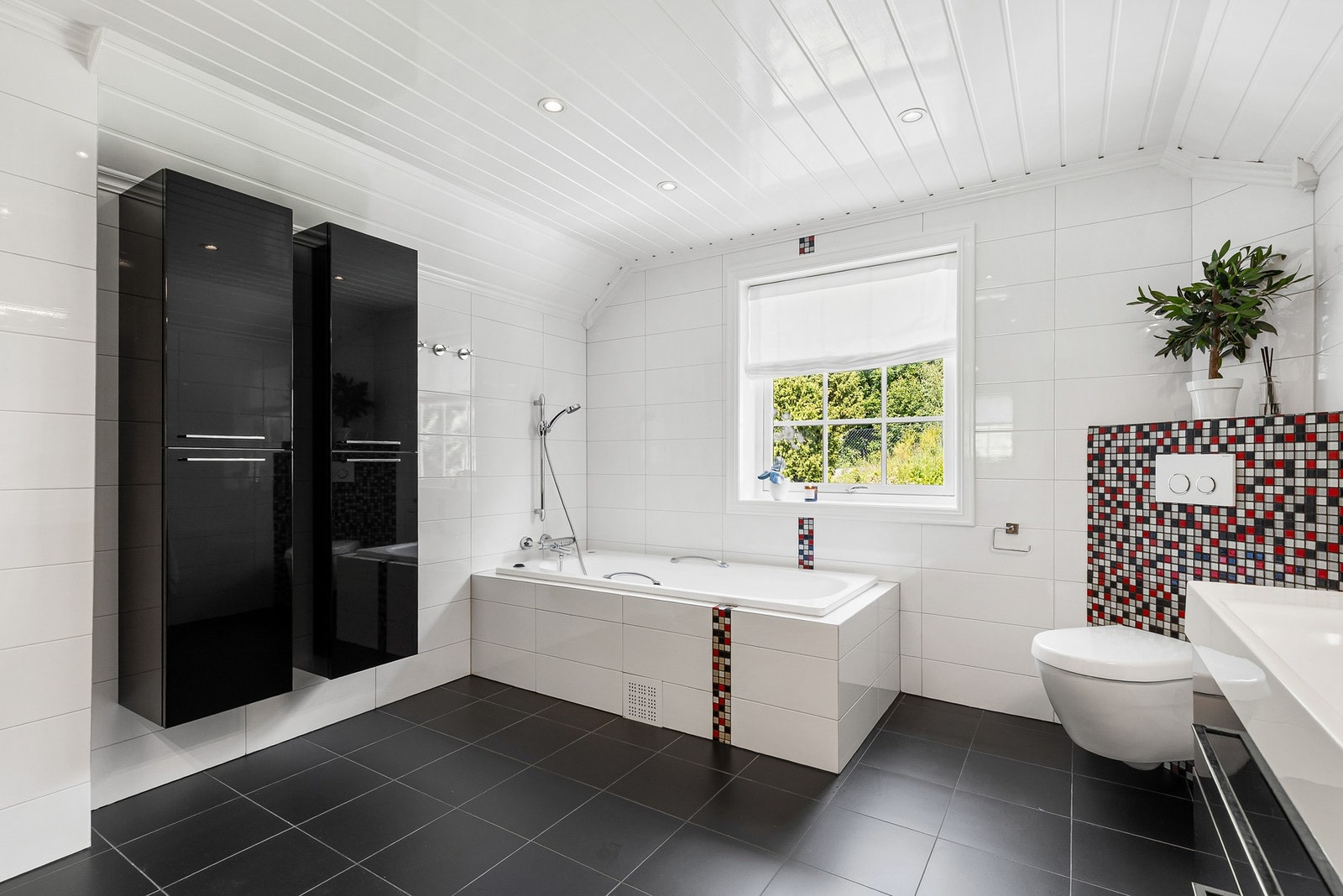 Badet er innredet med innebygget badekar, vegghengt toalett og dusjhjørne med innfellbare glassdører og veggmontert dusjarmatur Galleribilde