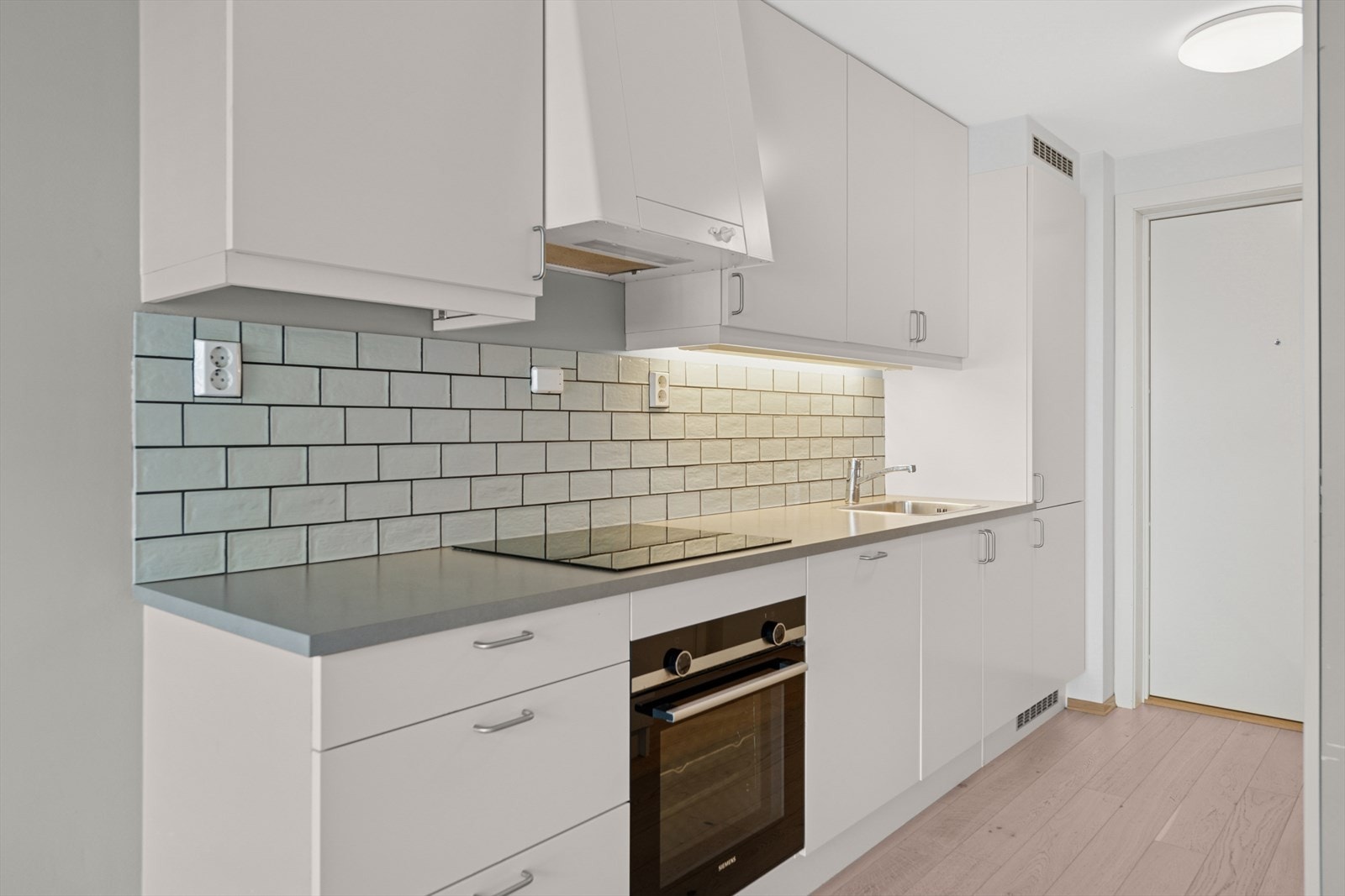 Kjøkkeninnredning fra 2018 med laminerte skrog, glatte fronter og laminat benkeplate med kjøkkenkum.
Kjøkkenet er videre utstyrt med kjøkkenventilator, samt fliser og belysning over benkeplate. Galleribilde