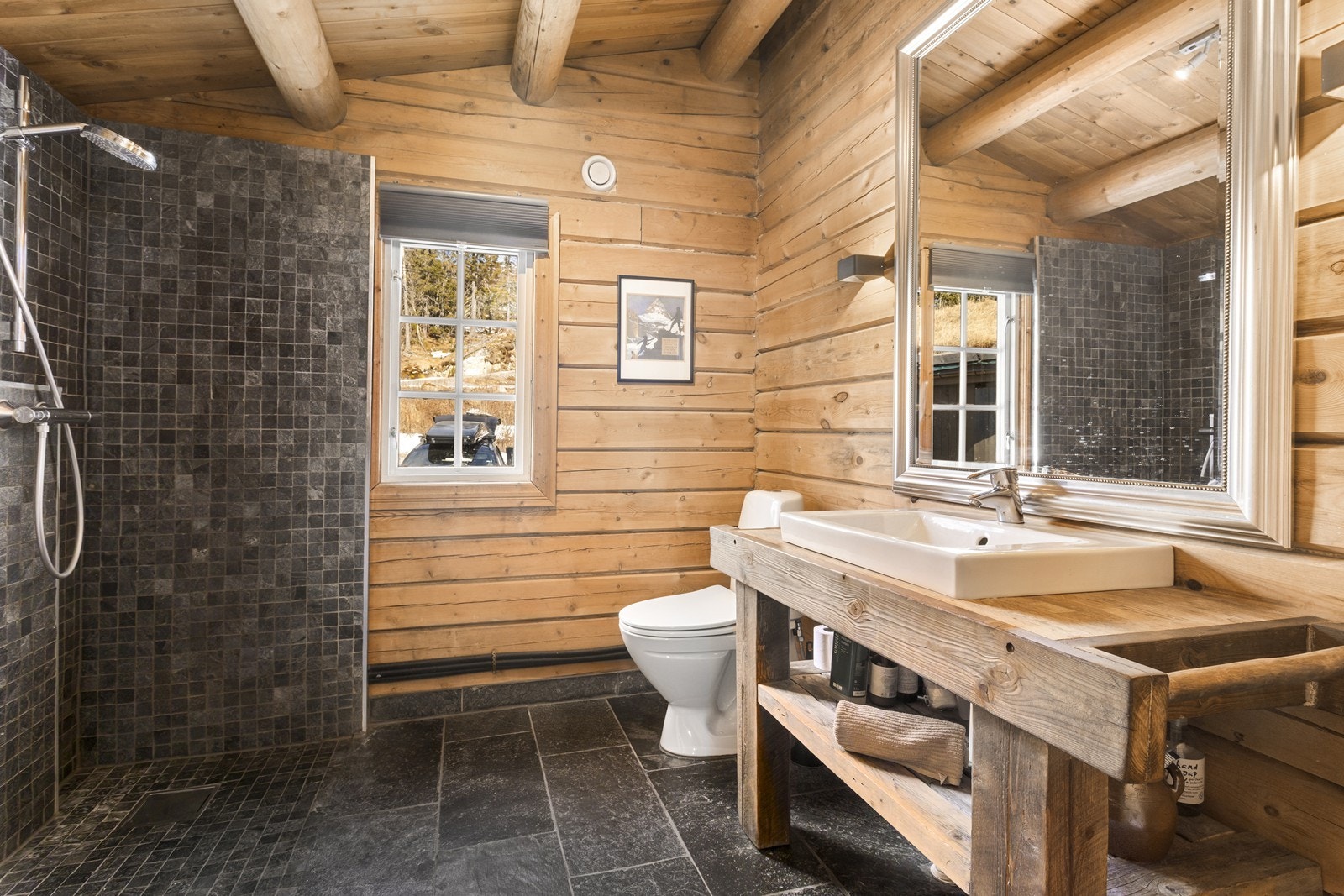 Innredet hytte bad med skiferfliser på gulv med varmekabler. I badet er det montert en rustikk innredning med nedfelt servant, frittstående toalett og åpen hjørnedusj. Opplegg for vaskemaskin. Galleribilde