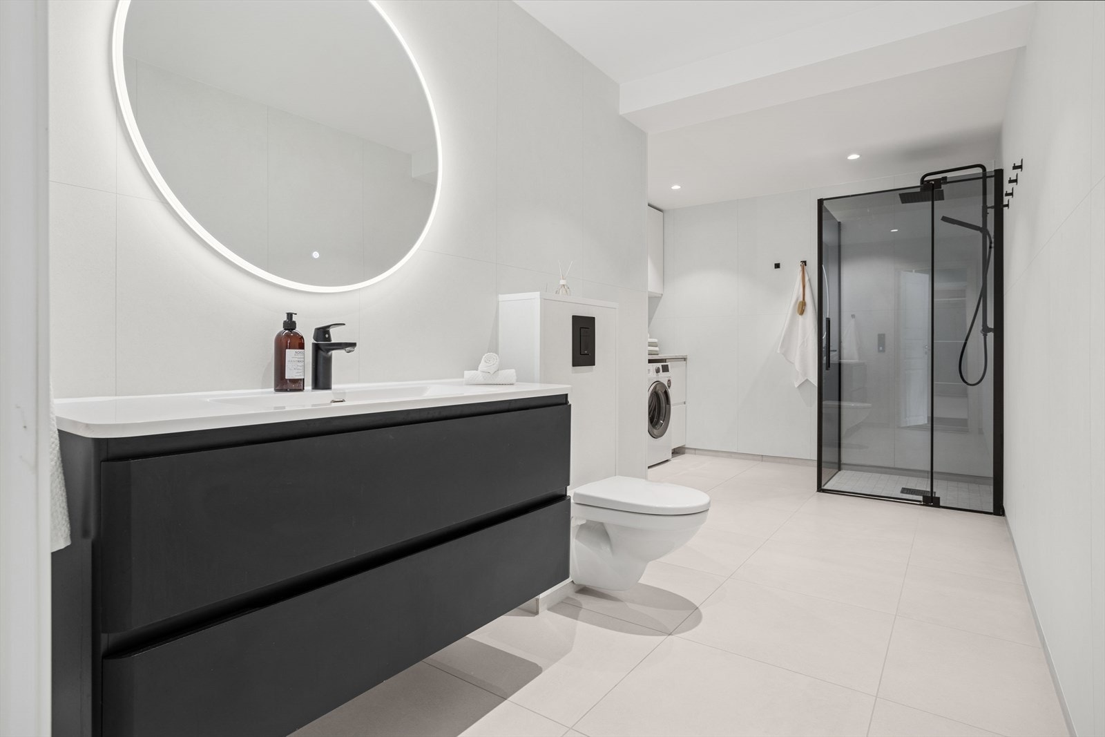 Badet inneholder dusjhjørne med glassdører og bunnlist, vegghengt toalett, servant med underskap, speil på vegg og opplegg for vaskemaskin/tørketrommel, samt vannfordelerskap og varmtvannsbereder Galleribilde