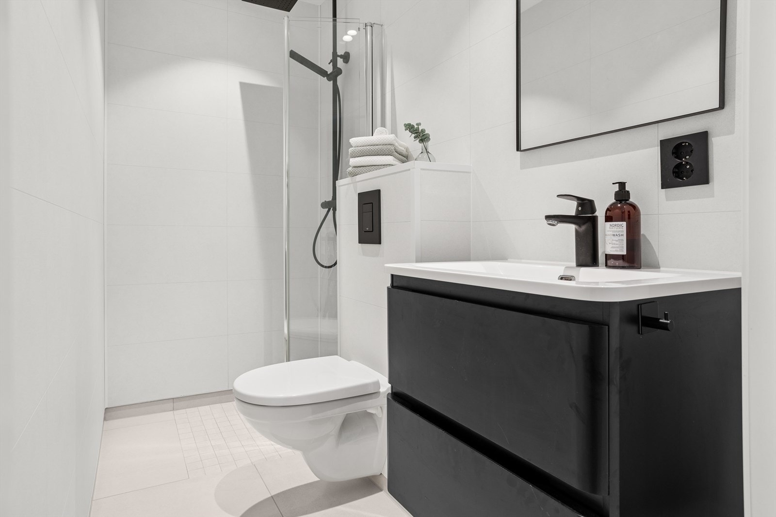 Bad 1: Lyst bad med fliser på gulv over nedstøpte, elektriske varmekabler. Badet inneholder dusjhjørne med innfellbare glassvegg, vegghengt toalett, servant med underskap og speil på vegg Galleribilde