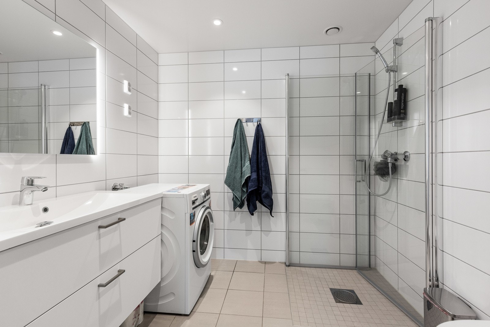 Delikat flislagt baderom med større servant med skuffer, dusjhjørne, veggmontert wc og opplegg for vaskemaskin/trommel. Galleribilde