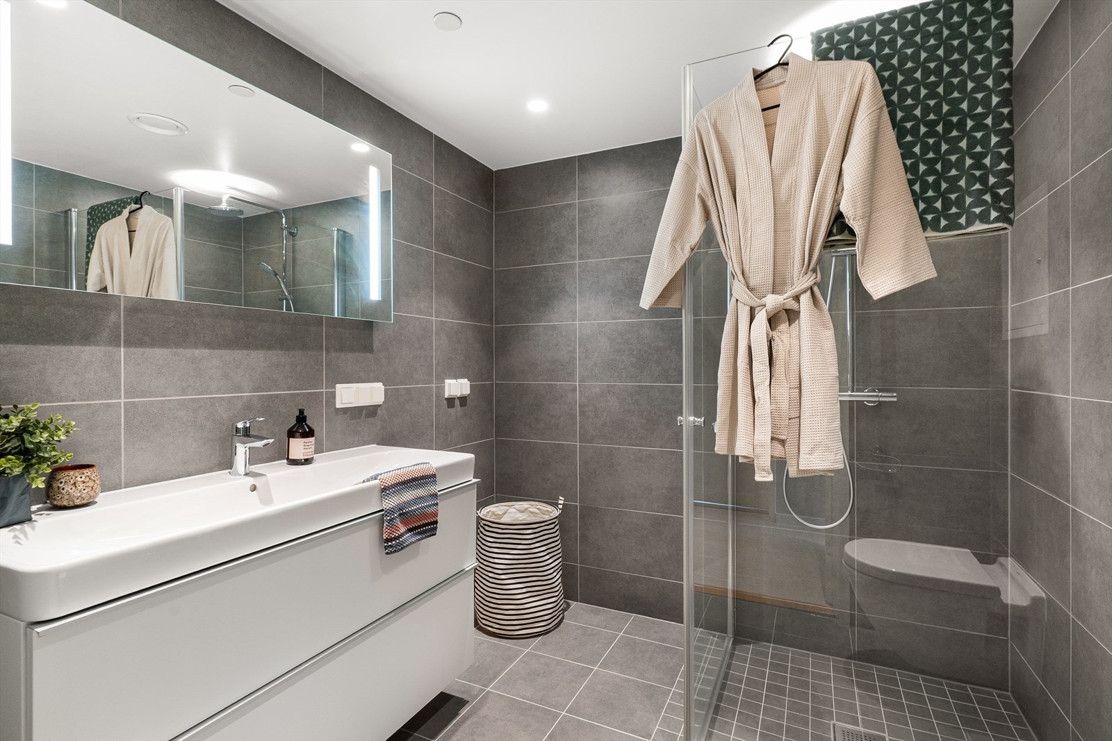 Badet har innredning med nedfelt servant, vegghengt toalett, vegghengt speil, dusjhjørne/vegger og opplegg for vaskemaskin. Galleribilde