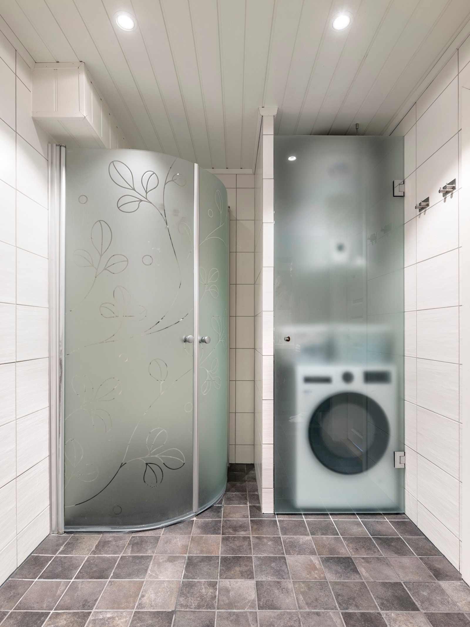 Badet i første etasje består av en heldekkende servant på brun skuffeseksjon med trestruktur, matchende høyskap og et dobbelt speilskap med belysning. Badet har også et veggmontert toalett og et dusjhjørne med innfellbare, frostede glassdører. Galleribilde