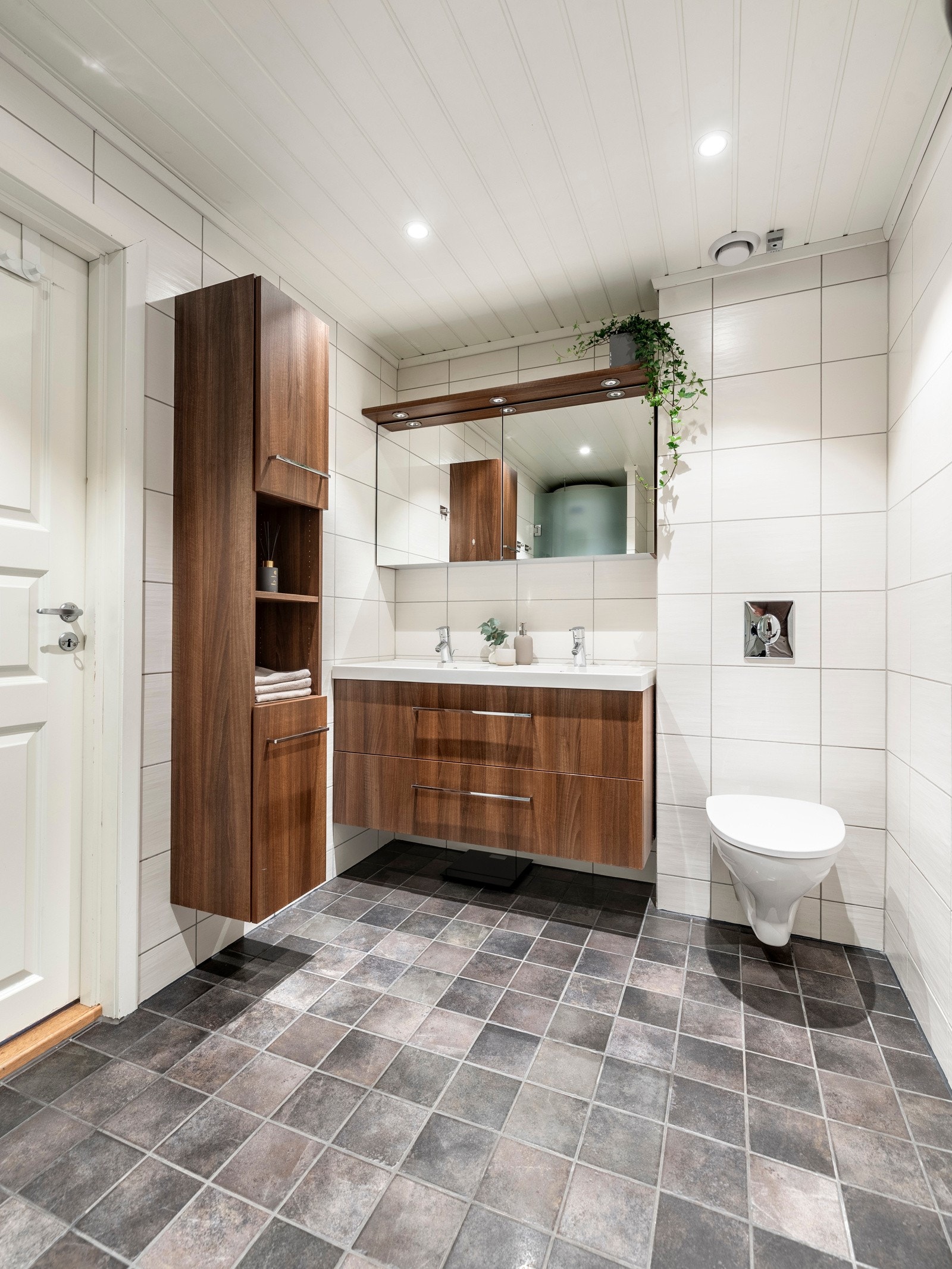 Badet i første etasje er fra 2010, et kombinert vaskerom. Opplegg for vaskemaskin er plassert i en egen nisje bak frostet glassdør. Rommet har grå fliser på gulv med gulvvarme, lyse fliser på veggene og hvitmalt panelhimling med downlights. Galleribilde