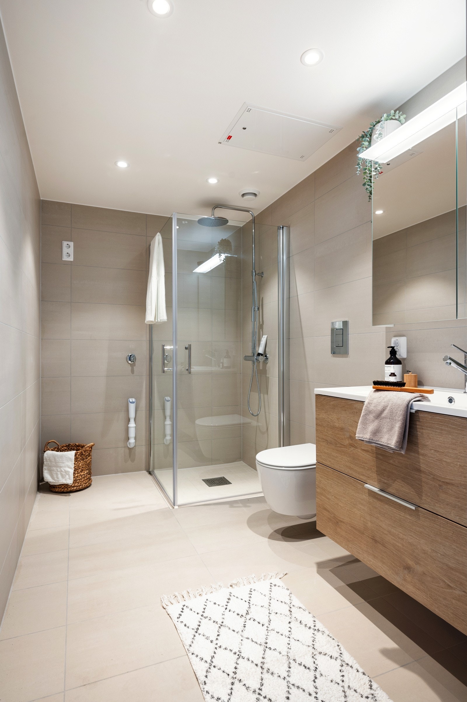 Et stilrent baderom med lune fliser og moderne innredning, hvor den nedsenkede dusjsonen er flislagt med matchende 5x5 cm fliser for et helhetlig preg, mens dusjdører med kromdetaljer setter prikken over i-en med sitt elegante uttrykk Galleribilde