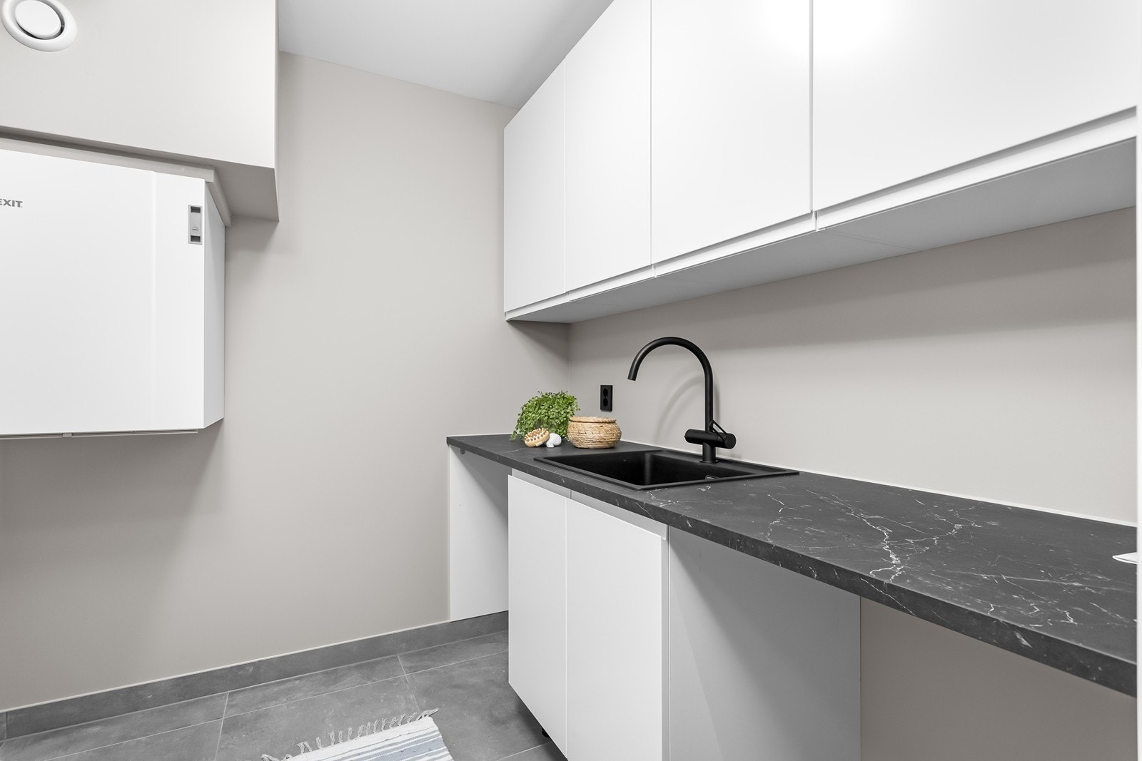 Praktisk vaskerom med oppbevaringsmuligheter i overskap, og en funksjonell benkeplate med nedfelt vask og avsatt plass til vaskemaskin og tørketrommel. Galleribilde