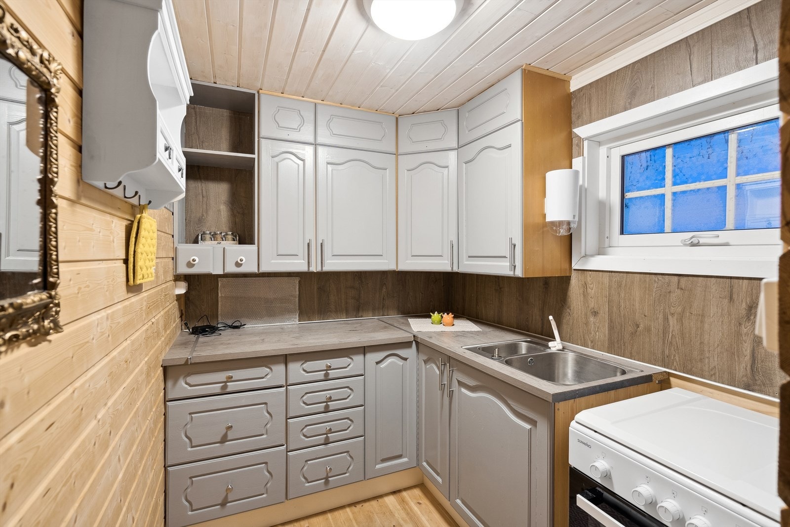 Kjøkkenet er praktisk plassert i egen sone og godt utnyttet med innredning i en L-vinkel. Kjøkkeninnredningen har malte, profilerte fronter, laminert benkeplate med dobbel vaskekum og frittstående gasskomfyr. Galleribilde