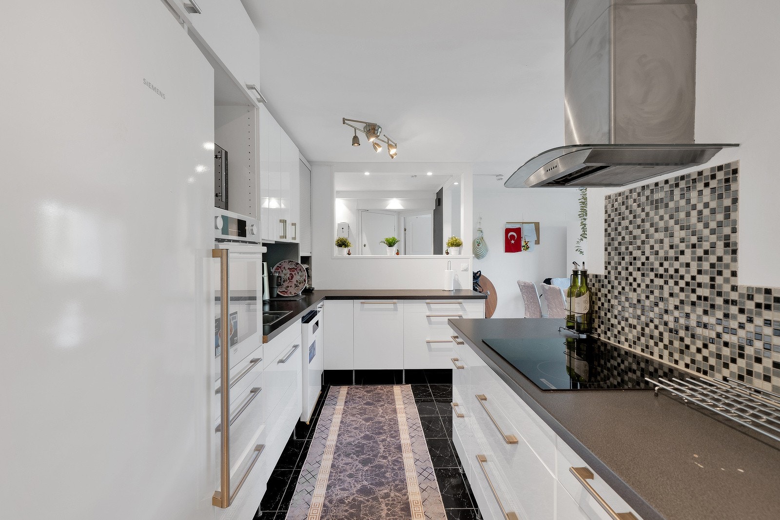 Innredningen består av glatte, høyglans hvite fronter med integrerte håndtak, overskap og høyskap som gir god lagringsplass. Benkeplater i mørk laminat gir en kontrastfylt og stilren effekt, og mellom benk og overskap er det montert mosaikkfliser i modern Galleribilde