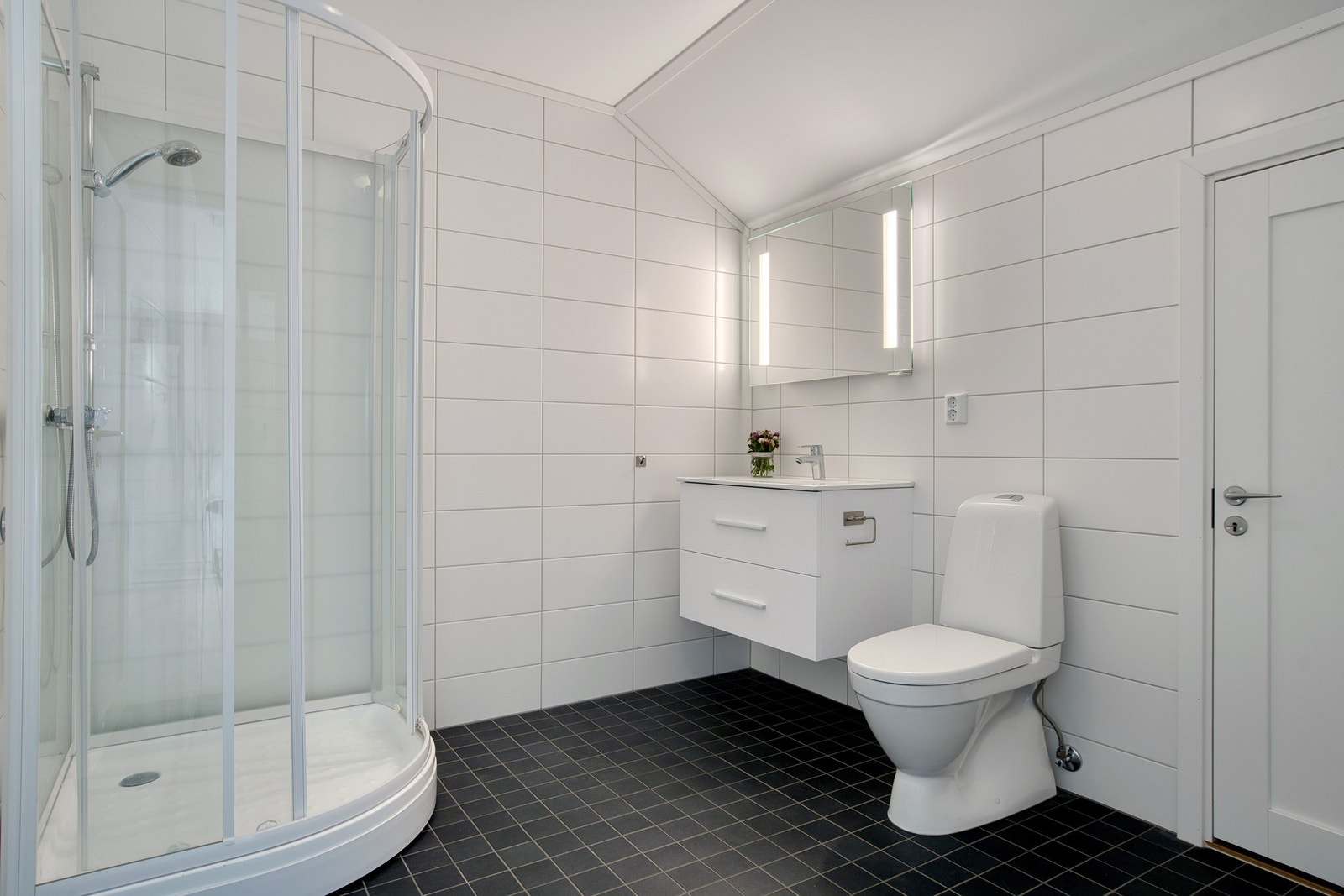 Badet er utstyrt med dusjkabinett, gulvstående wc, baderomsinnredning og speil med integrert belysning Galleribilde