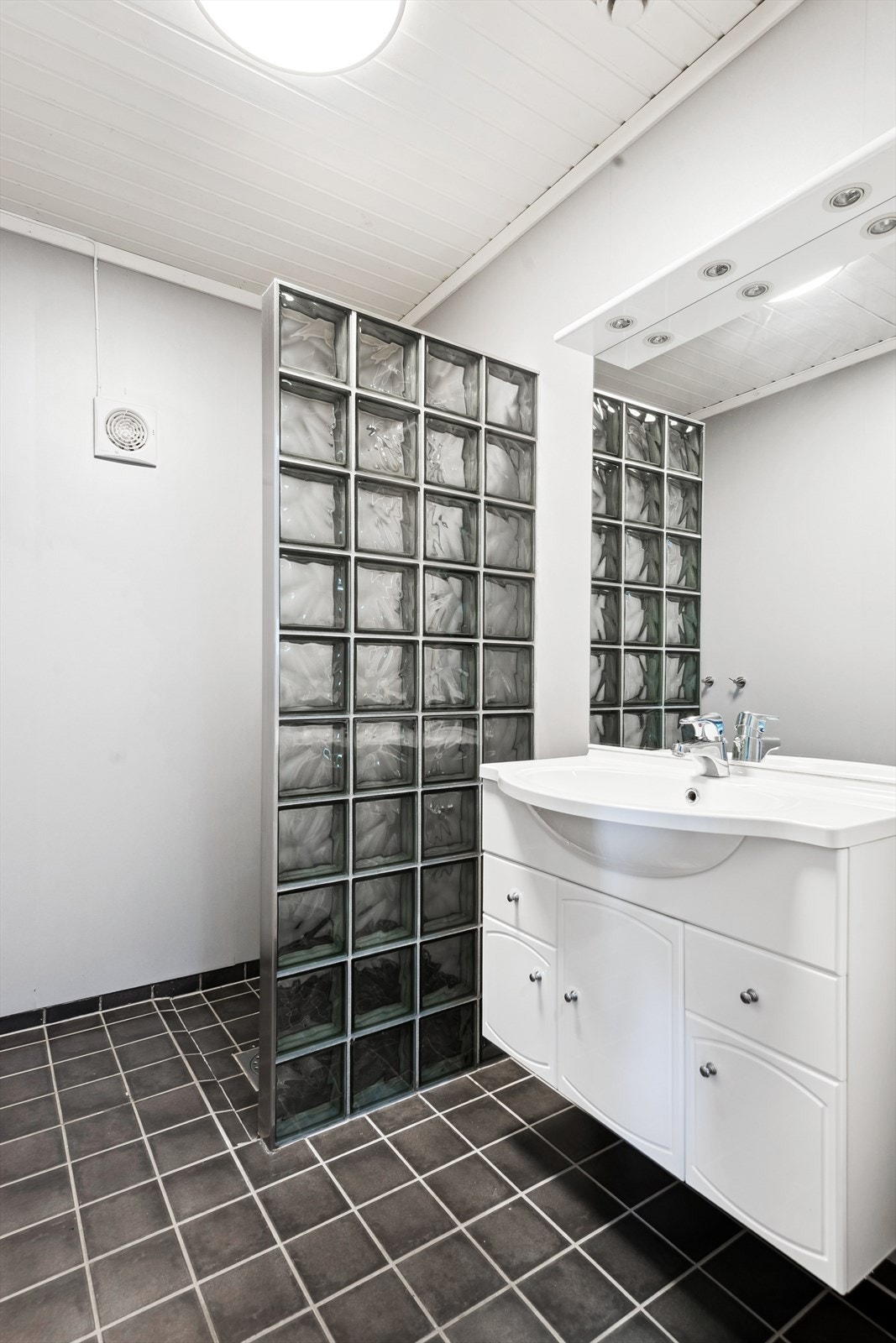 Rommet er utstyrt med servant i moderne baderomsinnredning med ettgreps blandebatteri, dusjkabinett og elektrisk vifte for effektiv ventilasjon. Tettesjikt med baderomspanel og plastsluk fra byggeåret gir trygghet og funksjonalitet. Galleribilde