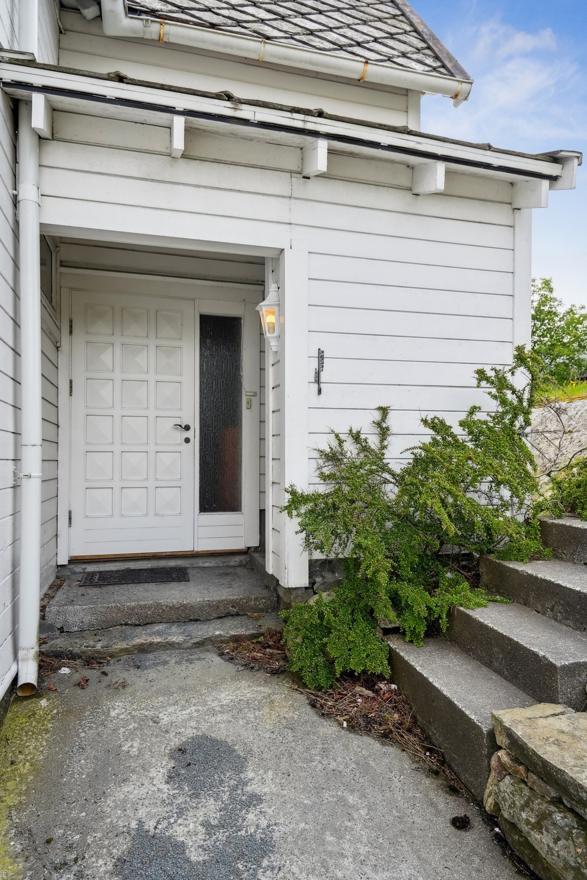 Inngangsparti på nordsiden med støpt dekke og takoverbygg. Det er montert belysning på veggen. Trapp opp til hage. Galleribilde