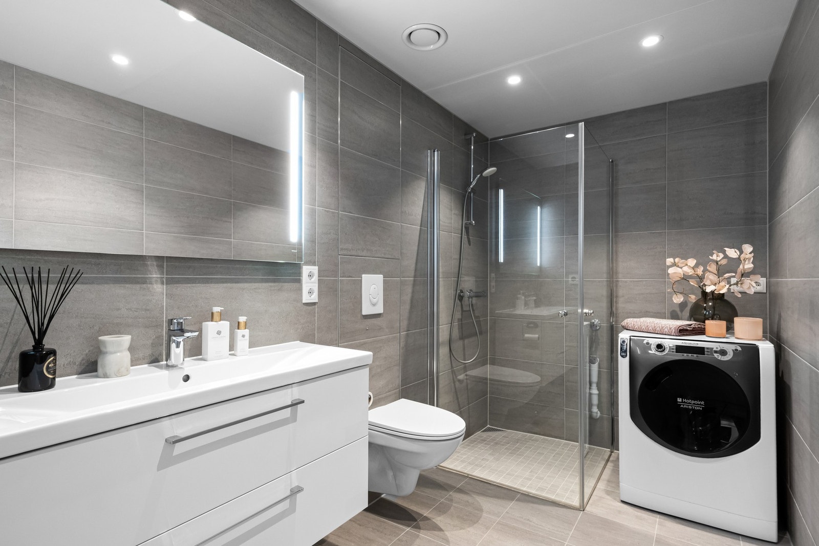 Badet er utstyrt med flislagte vegger og gulv, samt glatt himling. Det er montert dusjhjørne med svingdører i glass og opplegg for vaskemaskin / tørketrommel Galleribilde