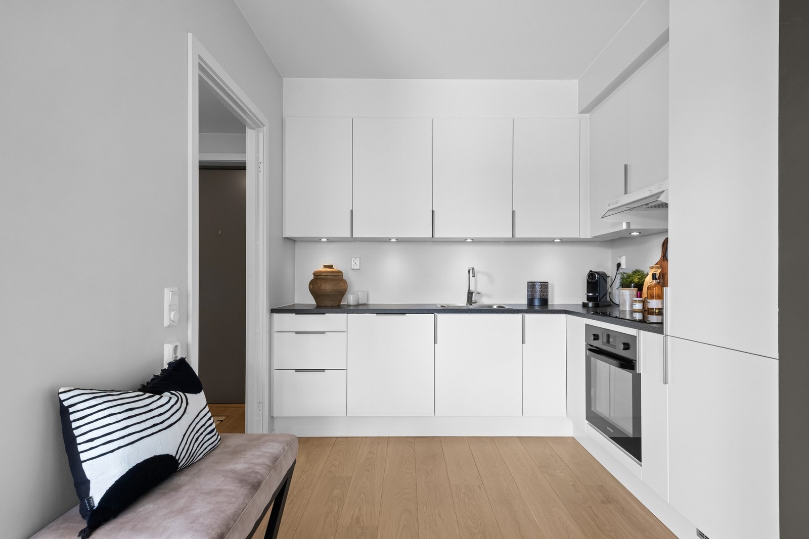 Kjøkkenet har en åpen løsning mot stuen og er utstyrt med glatte fronter og en laminat benkeplate med nedfelt kum i rustfritt stål Galleribilde