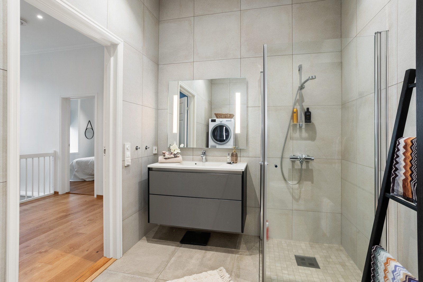 Lyst, pent og flislagt bad, med dusjhjørne, vegghengt toalett og servant med underskap og overspeil - Opplegg til vaskemaskin og tørketrommel. Galleribilde