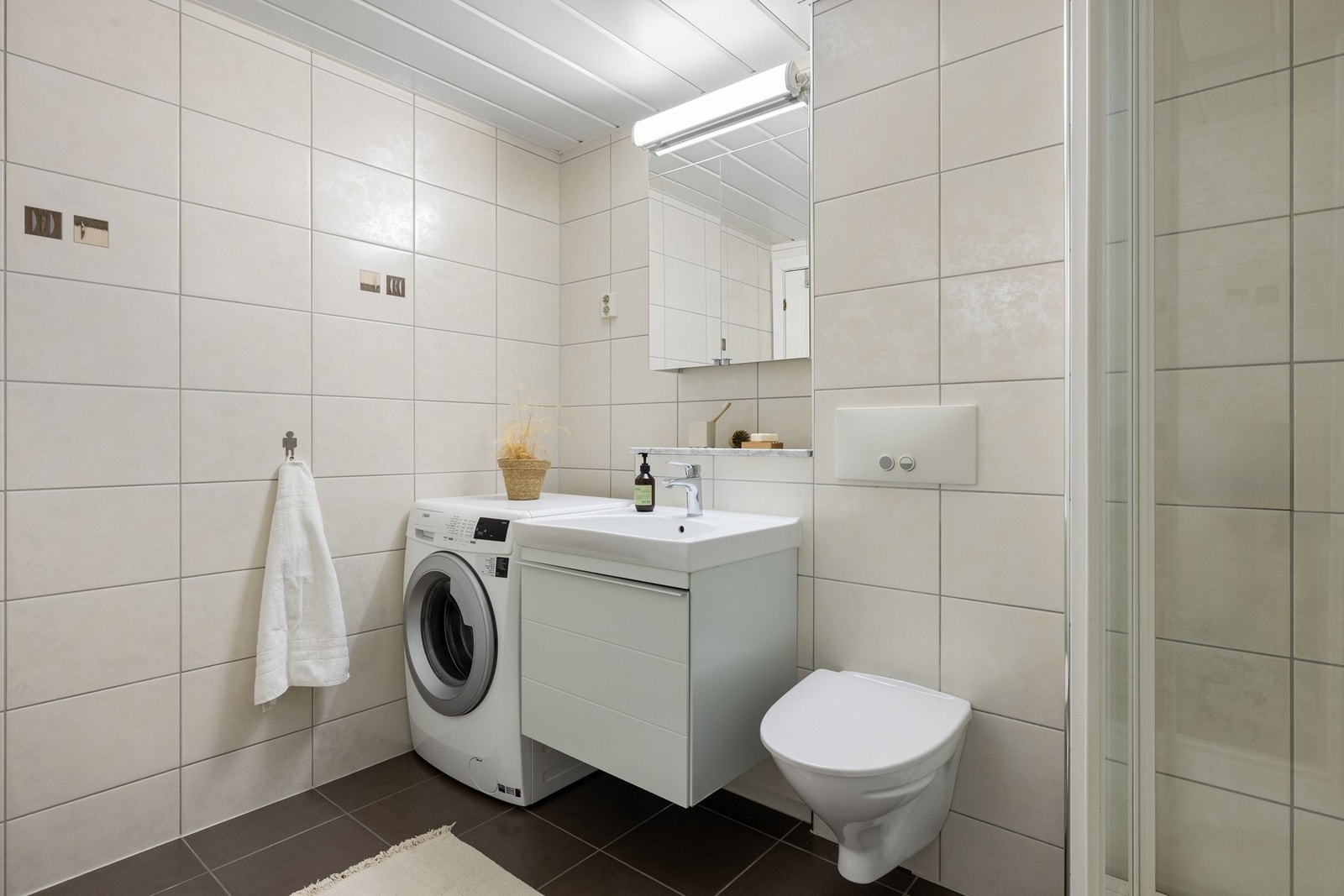 Badet er innredet med vegghengt toalett, opplegg for vaskemaskin, dusjkabinett og baderomsinnredning. Galleribilde