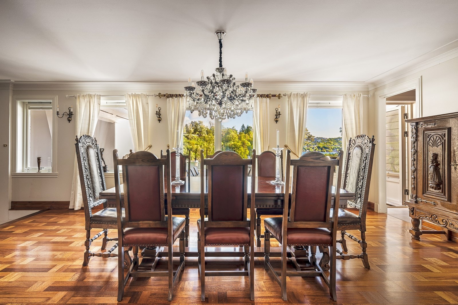Storslått spisestue med klassiske møbler og krystallkrone, omgitt av store vinduer som slipper inn rikelig med naturlig lys og gir vakker utsikt mot hagen og bukten. Elegant parkettgulv og gjennomført stil skaper en herskapelig atmosfære. Galleribilde