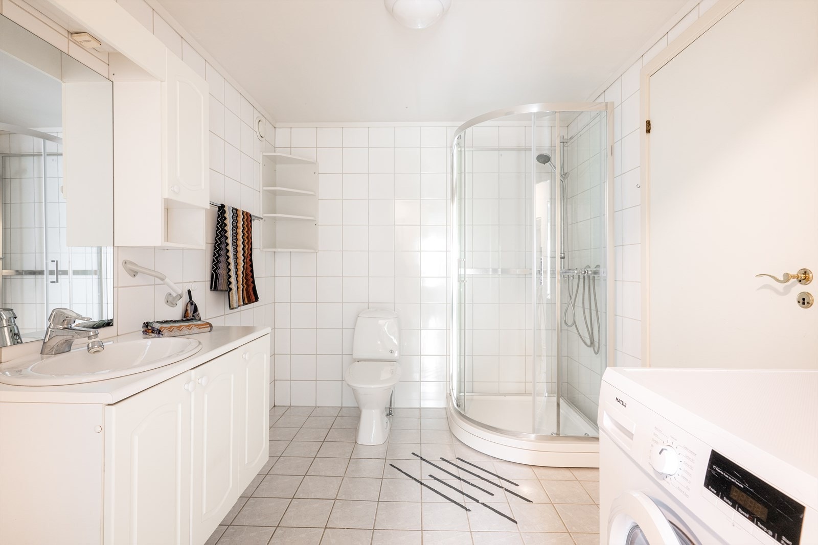 Flislagt bad med varmekabler i gulvet. Badet er utstyrt med toalett, dusjkabinett og servantinnredning med hvite, profilerte fronter. Det er også opplegg for vaskemaskin. Galleribilde