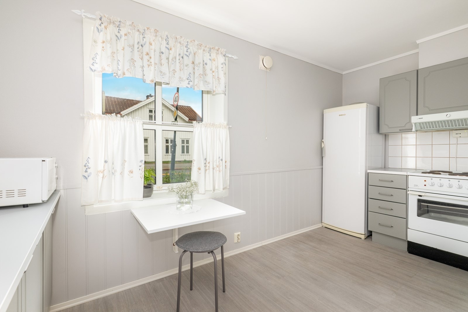 Koselig kjøkken med plass til et lite kjøkkenbord ved vinduet. Gulvet har laminat, og veggene er kledd med malt glassfiberstrie og brystningspanel, mens taket består av malt pusset betong. Galleribilde