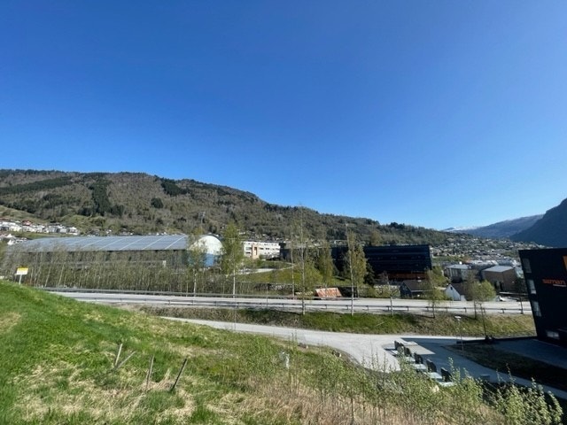 tomta med riksvegen og Campus Fosshaugane - Storhallen - Høgskulen i bakgrunnen. Kort veg til campus med bru over elva. Galleribilde