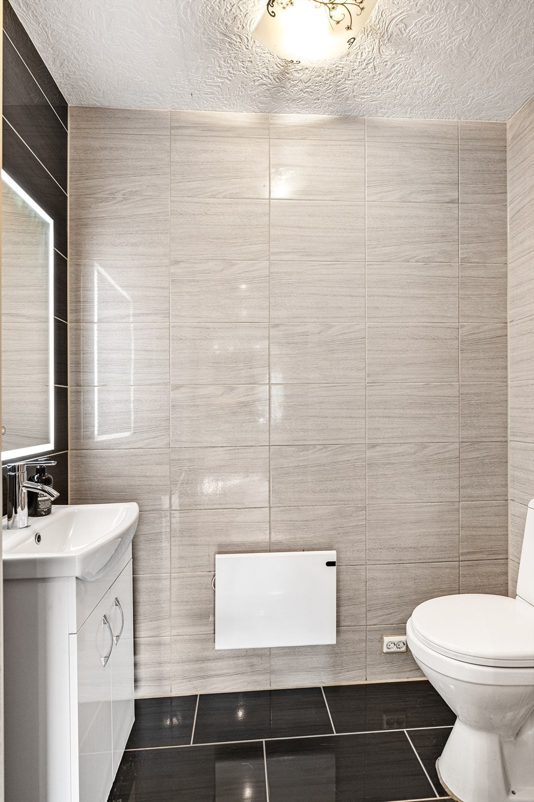 Toalettrommet har flis på gulv og vegger. Rommet inneholder servant med servantskap, vegghengt speil og toalett. Hvitmalte plater i tak Galleribilde