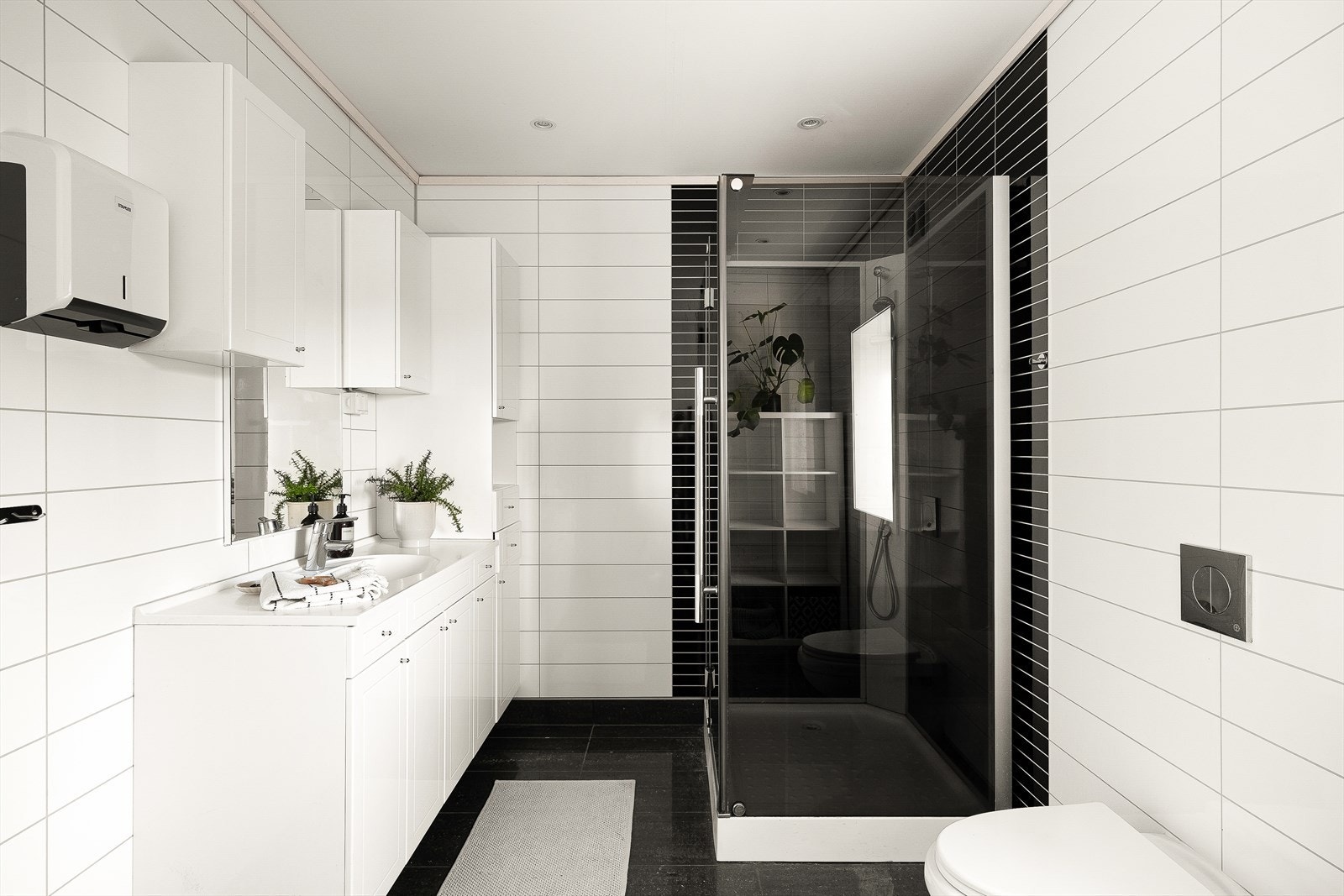 Badet i første etasje har mørke gulvfliser med varmekabler og våtromsplater på veggene. Det er innredet med dusjkabinett, servant med seksjon, langskap, overskap, speil og vegghengt toalett. Galleribilde