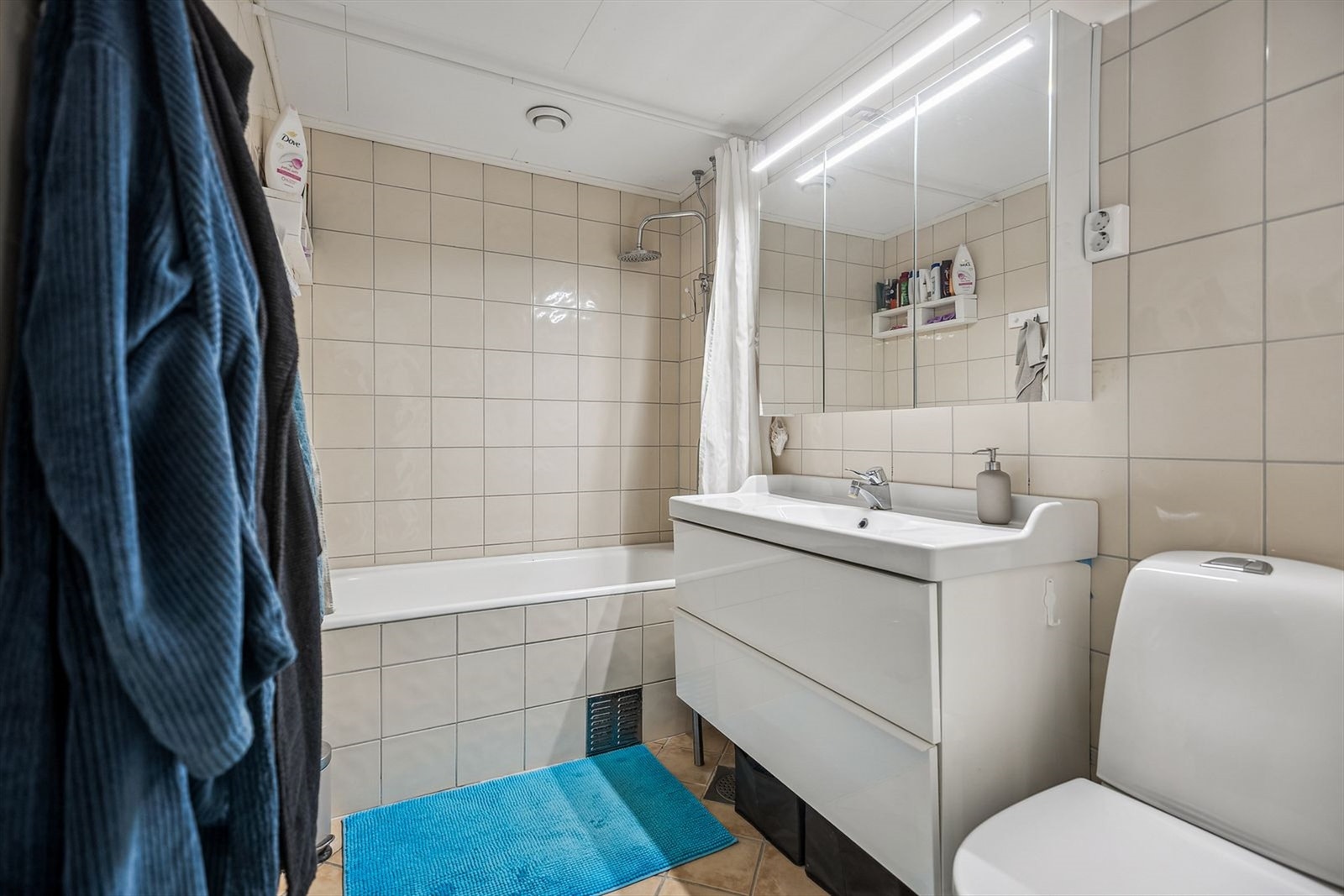 Badet er av god størrelse med flislagte flater, badekar, stor servant og et speilskap med belysning Galleribilde