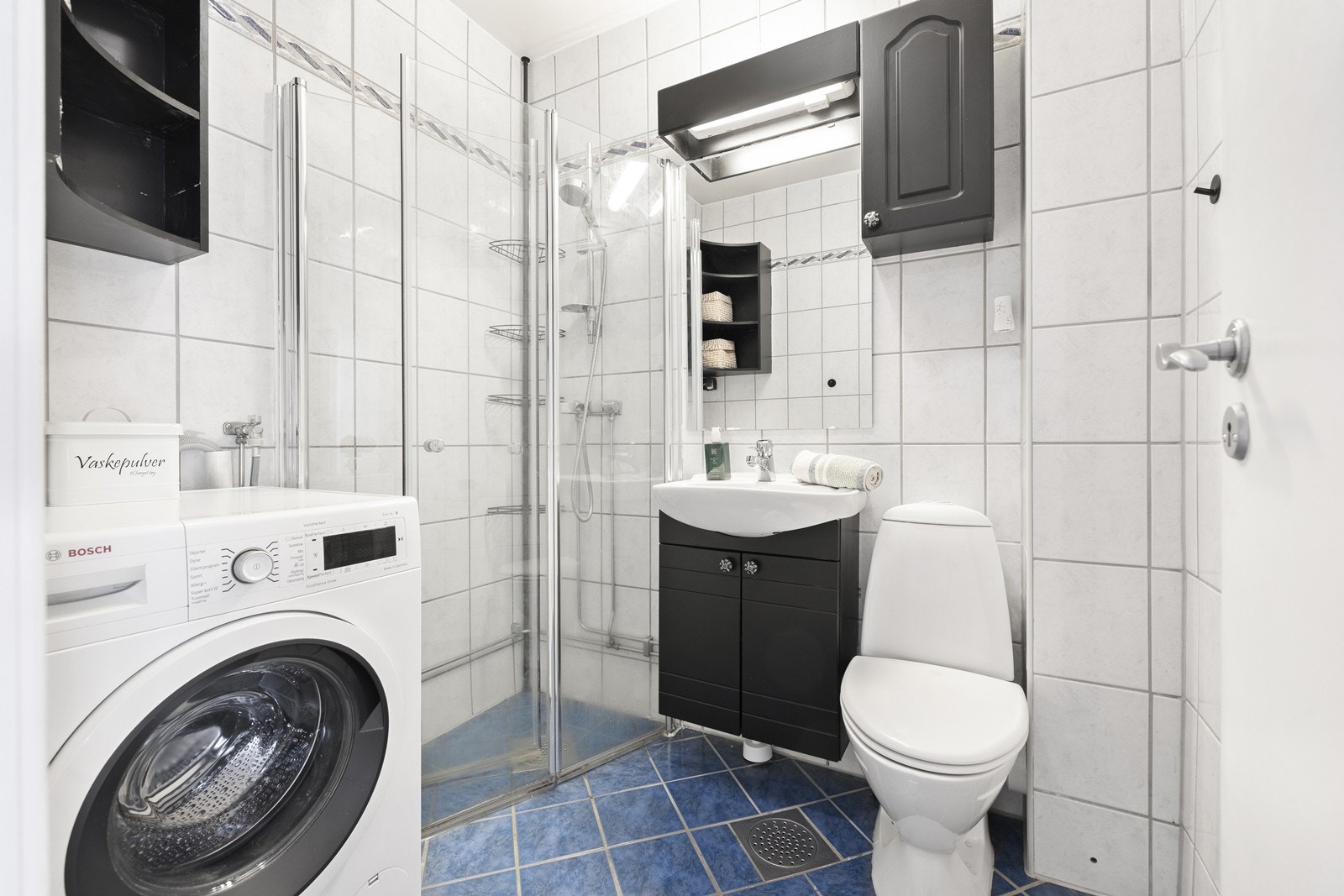 Flislagt bad med varmekabler i gulv, dusjhjørne med glassdører, opplegg for vaskemaskin og servant med underskap og overskap med speil, samt gulvstående toalett. Galleribilde