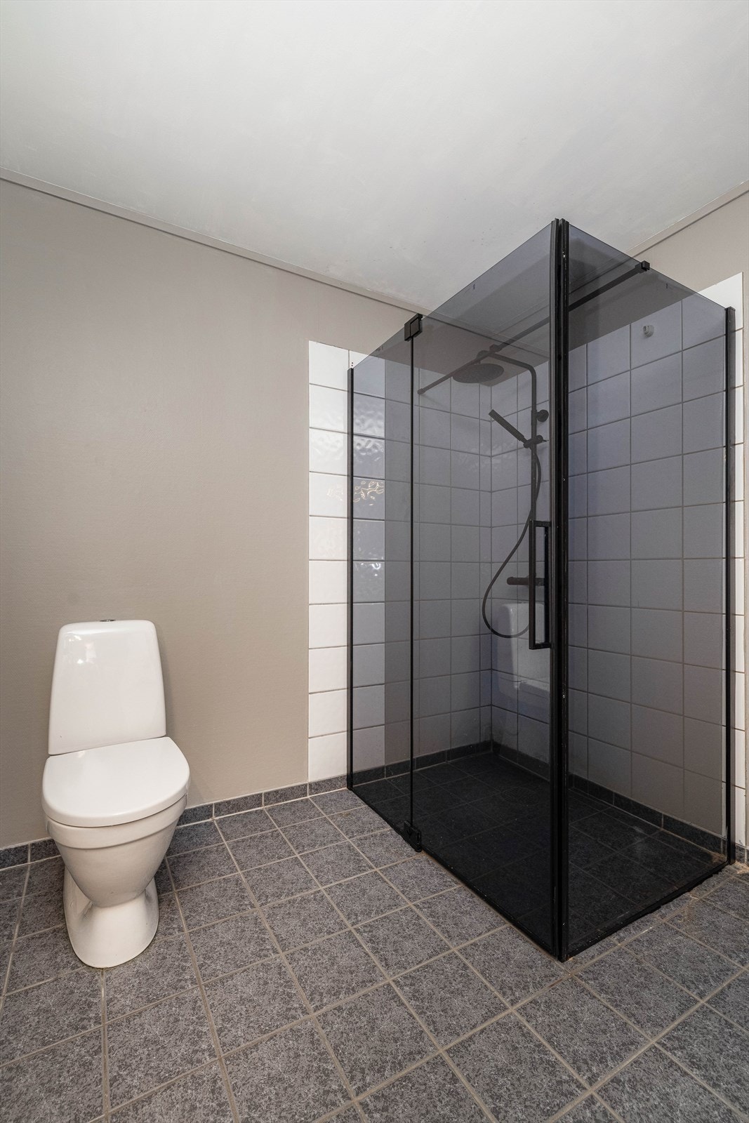 Fin dusjnisje med armatur på vegg og dusjdører i sort fargetone. Gulvmontert toalett. Galleribilde