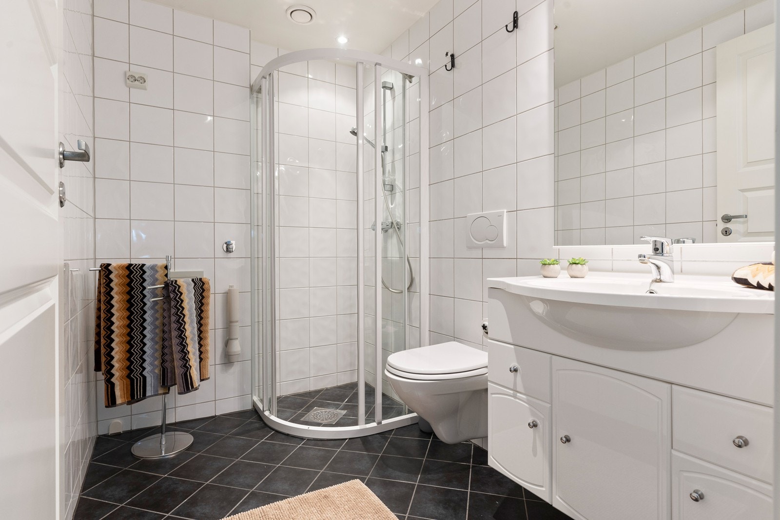 Pent flislagt baderom i tidløse fargetoner. Badet er utstyrt med vegghengt toalett, servant med skap og dusjnisje med kabinett. Galleribilde