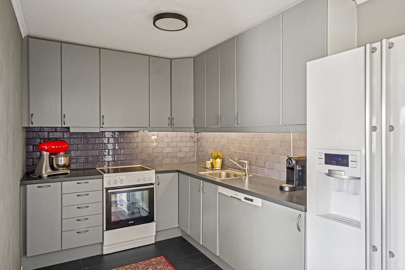Kjøkkeninnredning med glatte fronter, belysning under overskap, fliser på vegg over benkeskap. Laminat benkeplate med nedfelt kum i rustfritt stål. Galleribilde