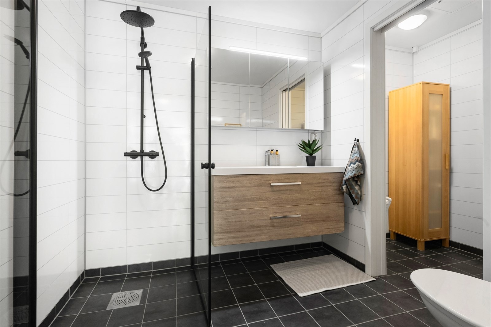 Badet er innredet med dusjløsning, servant og vegghengt toalett. Her er det flislagt gulv, baderomsplater på vegg og malt tak. Galleribilde