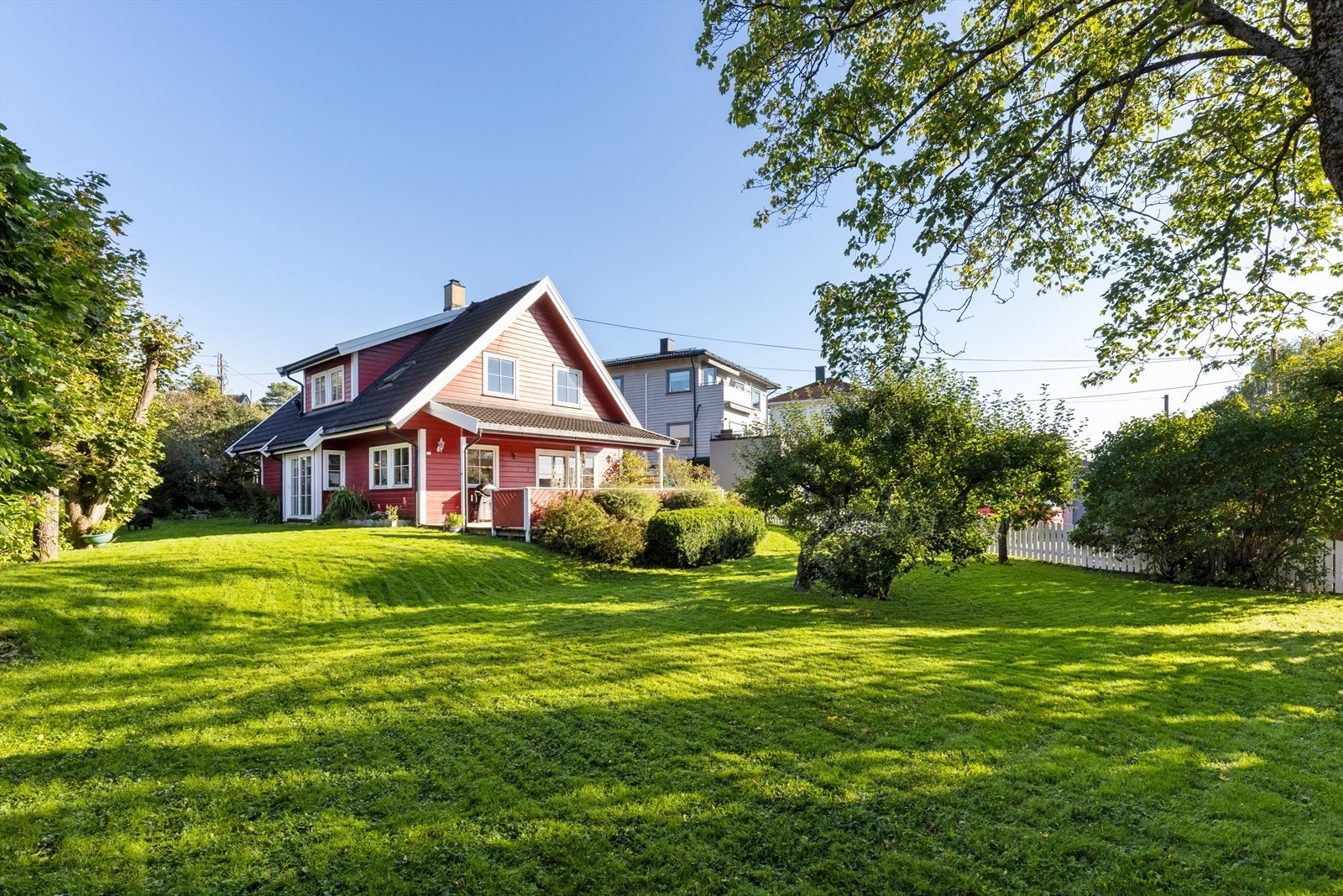 Boligen ligger omkranset av frodige trær, busker og fargerik beplantning. Galleribilde