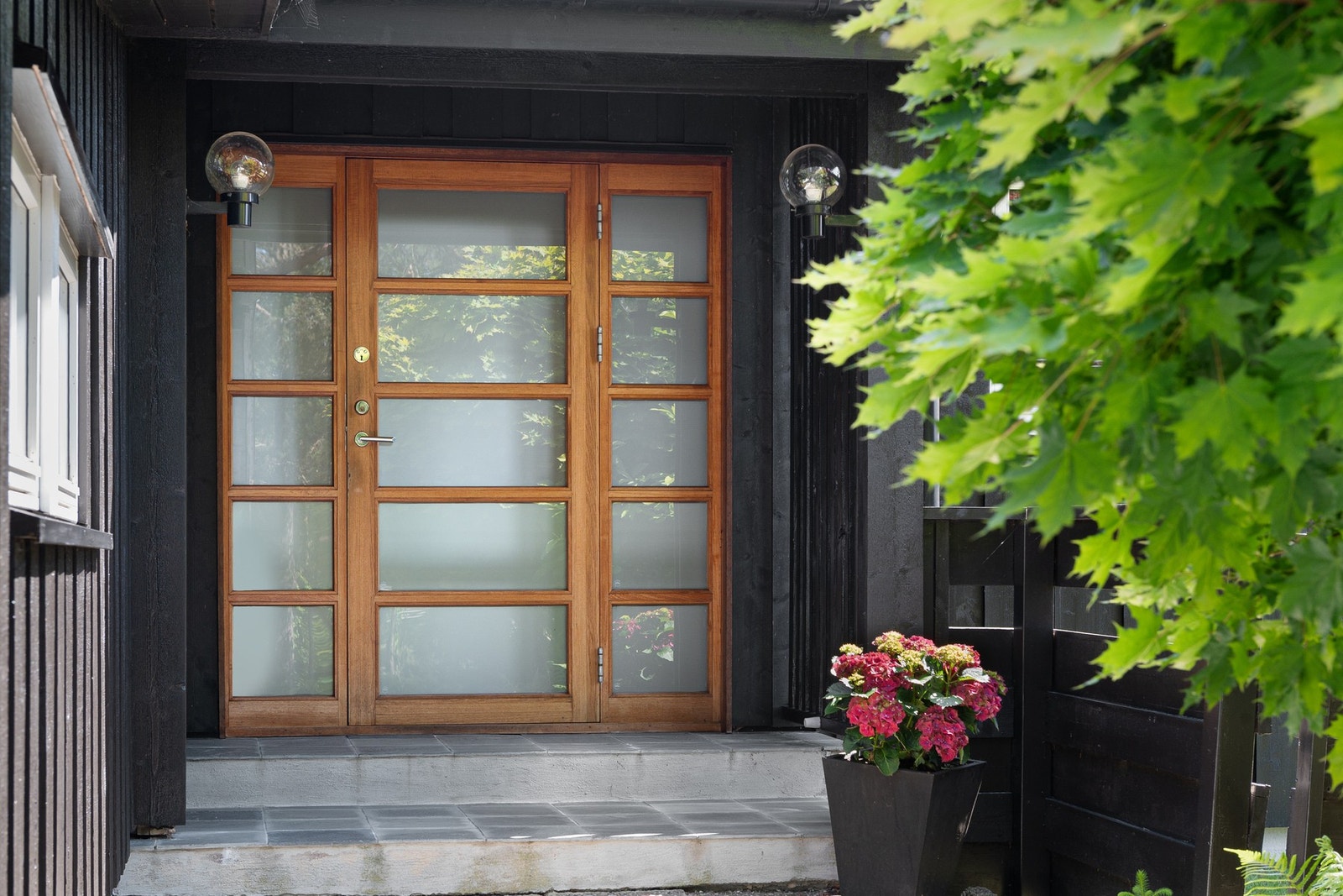 Inngangsparti med dobbel dør i tre og frostet glass, flankert av utelamper og blomsterdekorasjon. Ytterdør i teak. (Spes laget Østby trevare) Galleribilde