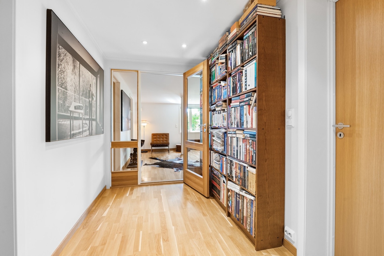 Lys gang med parkettgulv, bokhylle og speildør som leder inn til stue. Galleribilde