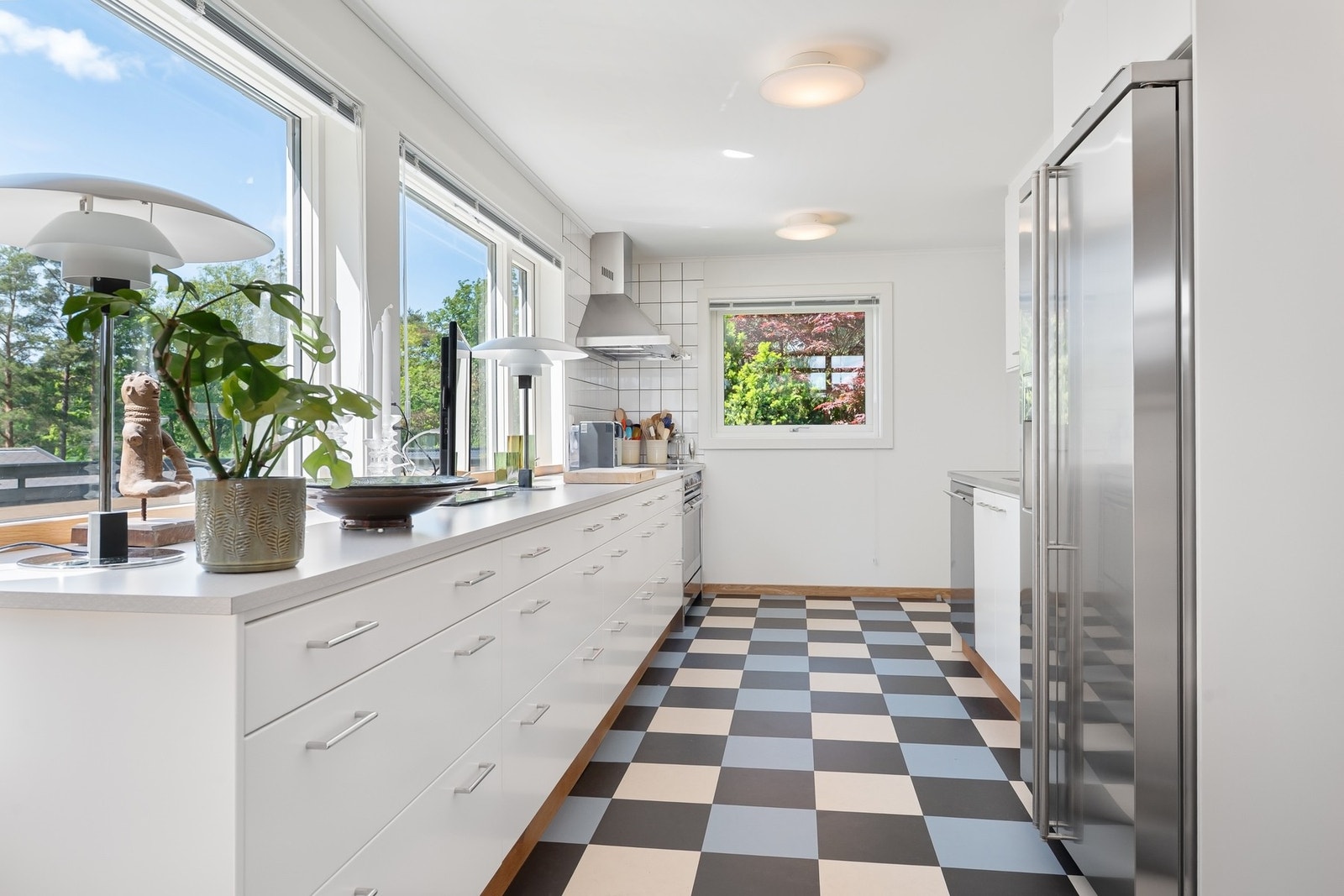 Lyst kjøkken med store vinduer, moderne hvitevarer og sjakkrutete gulv. Galleribilde