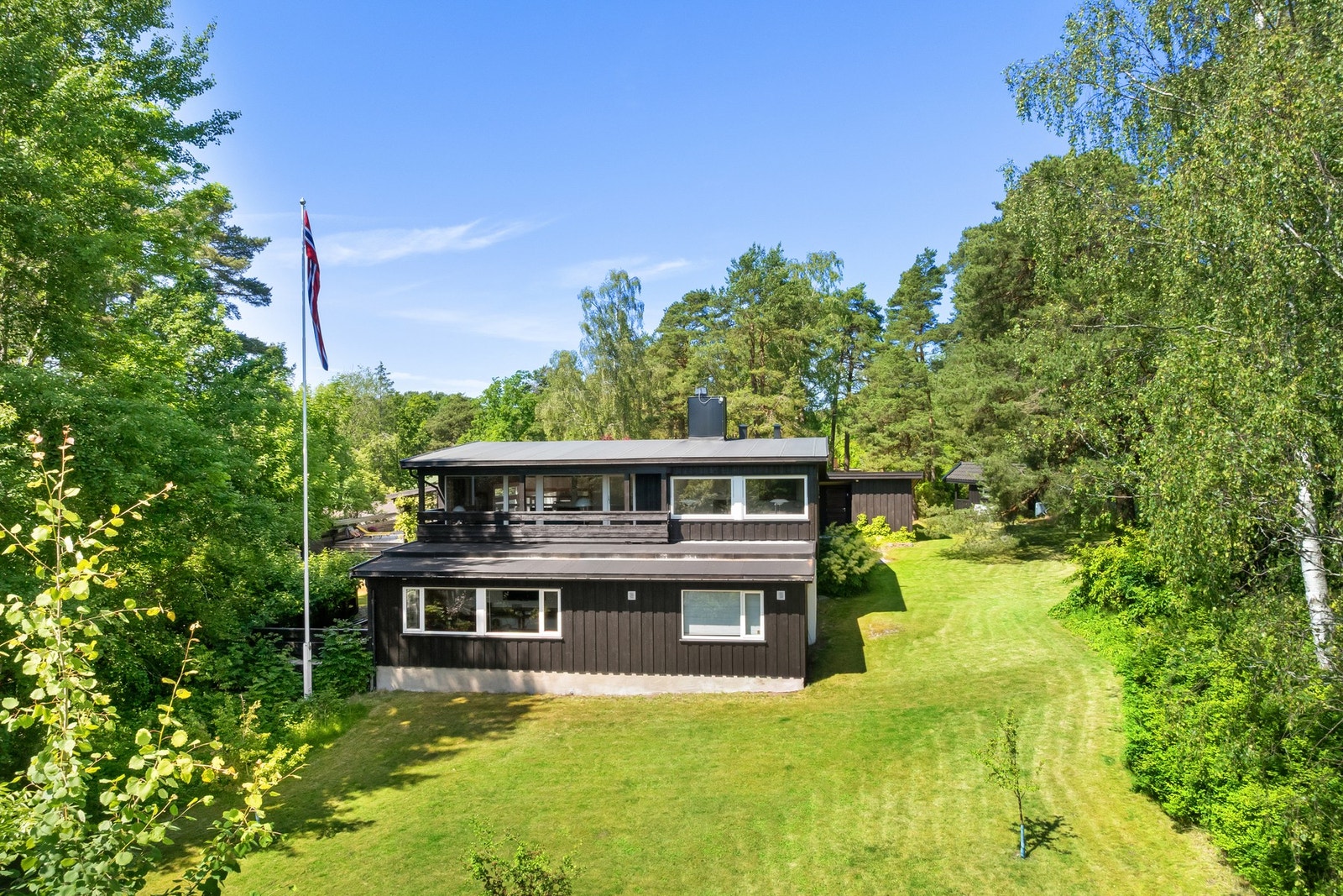 Moderne bolig med stor hage og flaggstang, omgitt av frodig vegetasjon og skog. Galleribilde