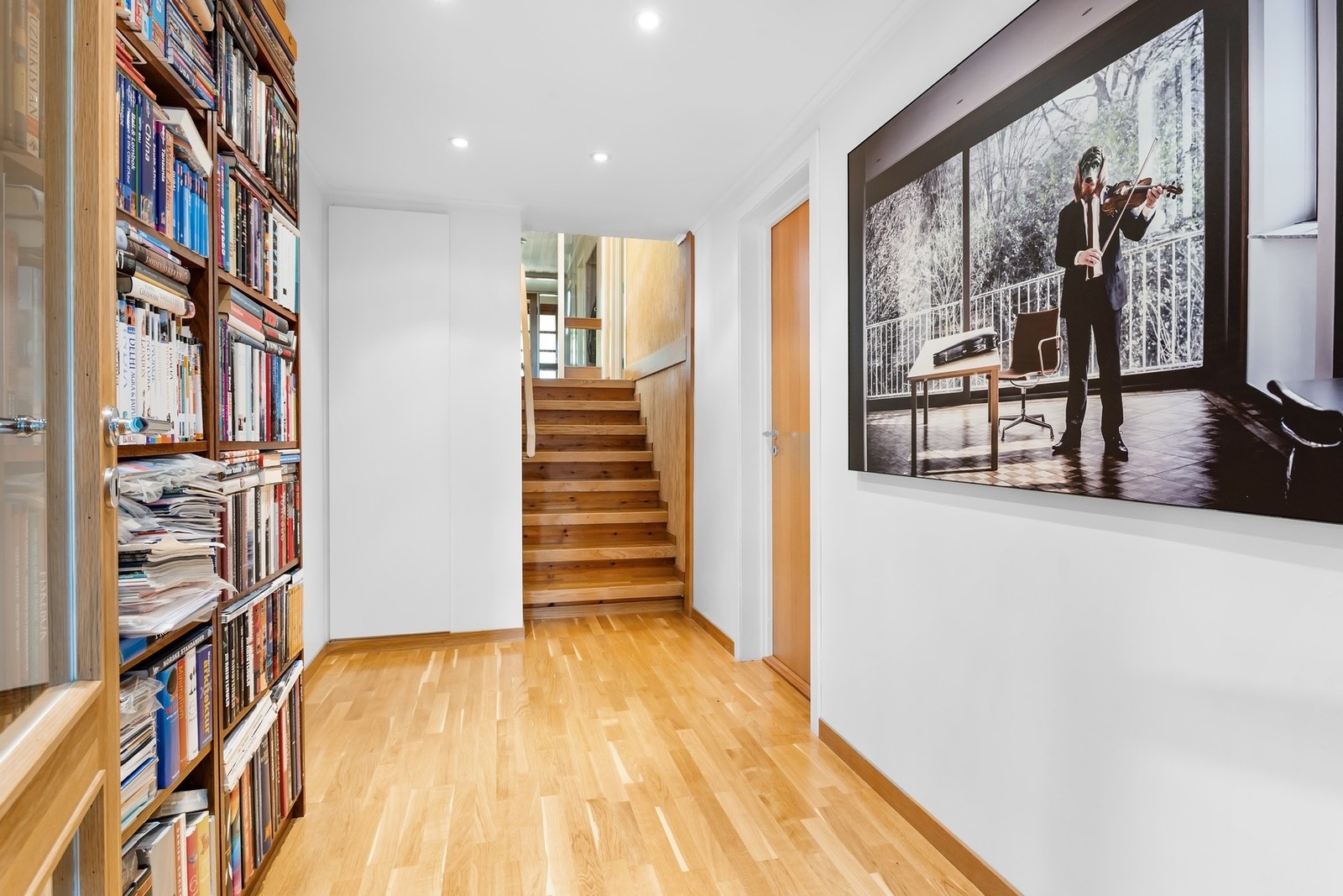 Lys gang med parkettgulv, bokhyller og trapp opp til neste etasje. Galleribilde