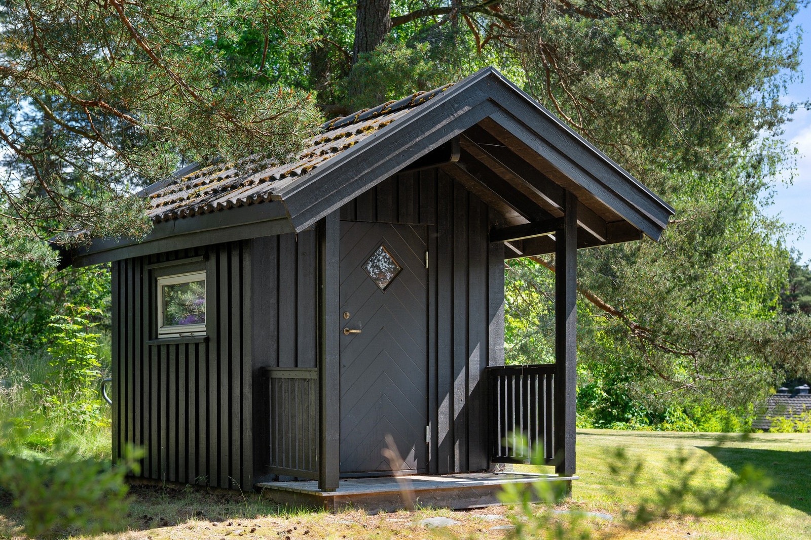 Liten, svartmalt hytte med skråtak og enkel inngangsdør, omkranset av trær og grøntområder. Galleribilde