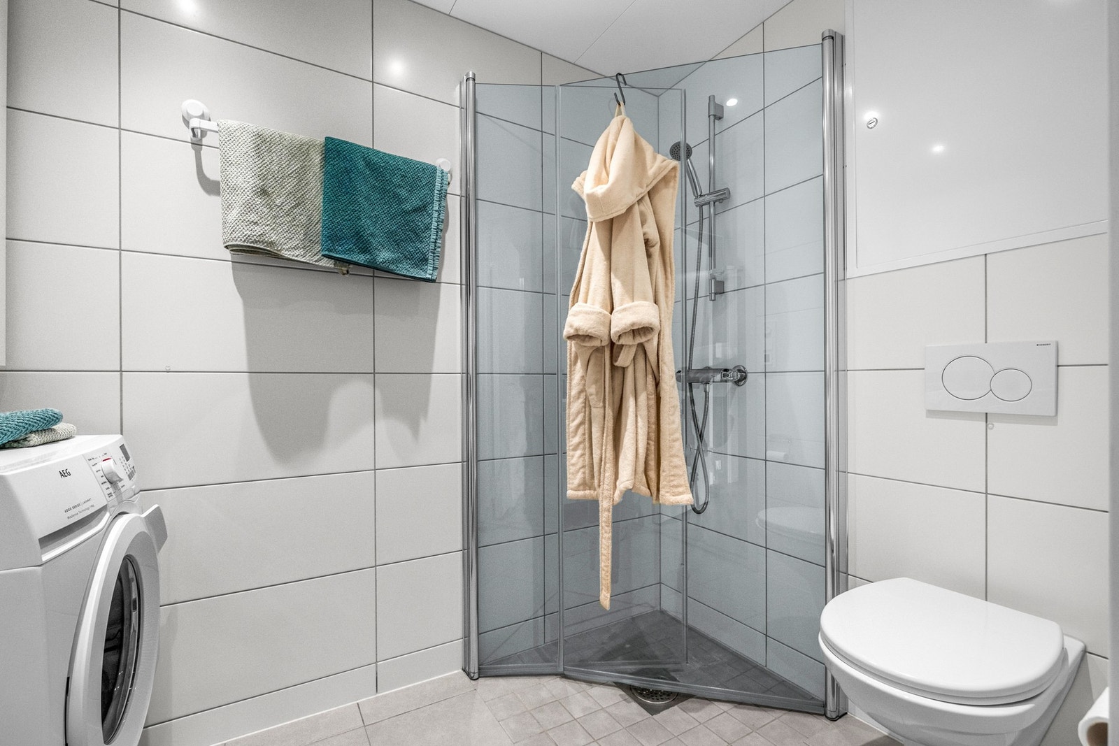 Badet er utstyrt med veggmontert toalett, dusjhjørnet med innfellbare glassdører og opplegg for vaskemaskin Galleribilde