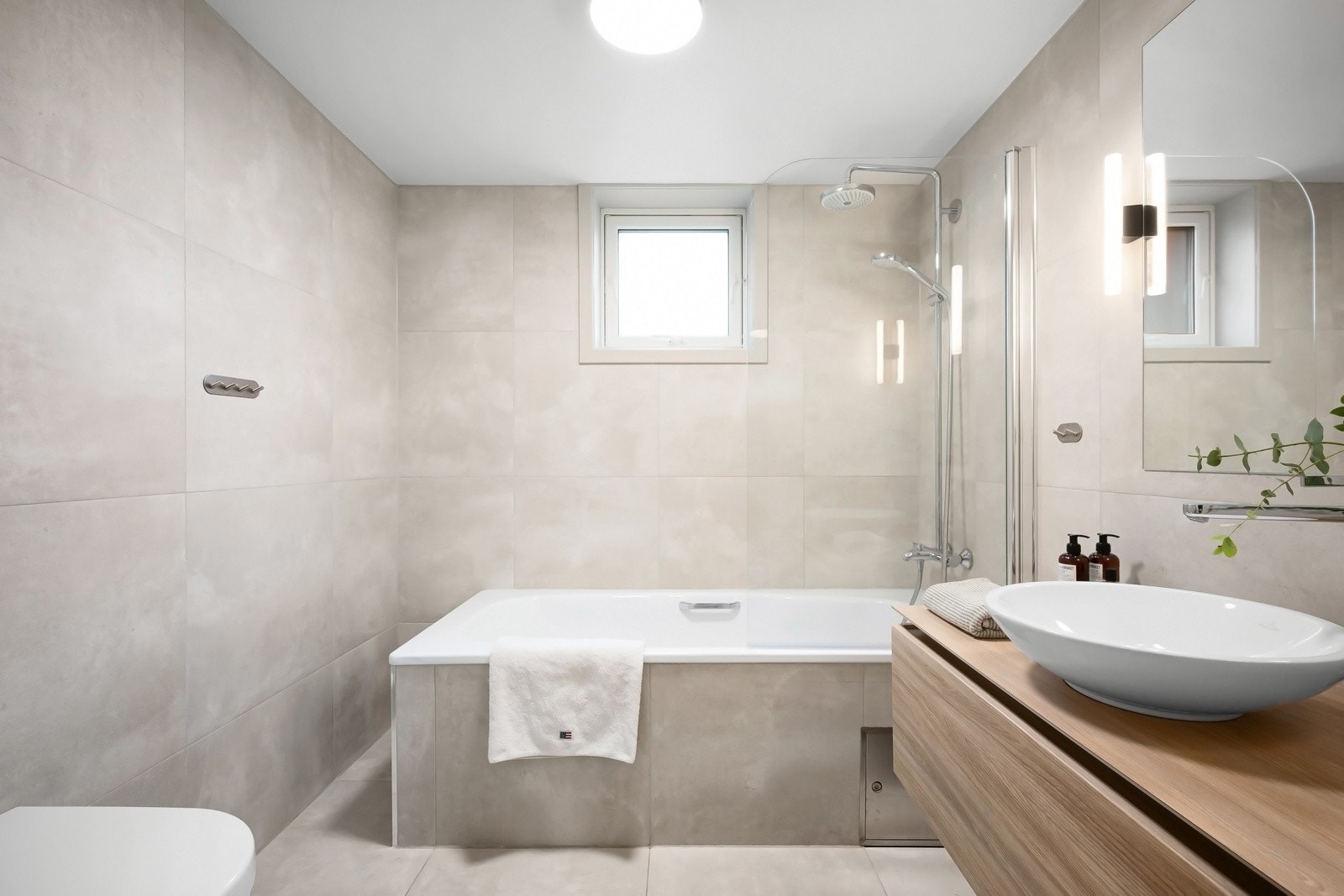 Stilrent baderom utstyrt med innfliset badekar med regnfallsdusj og håndholdt dusj samt veggmontert klosett. Galleribilde