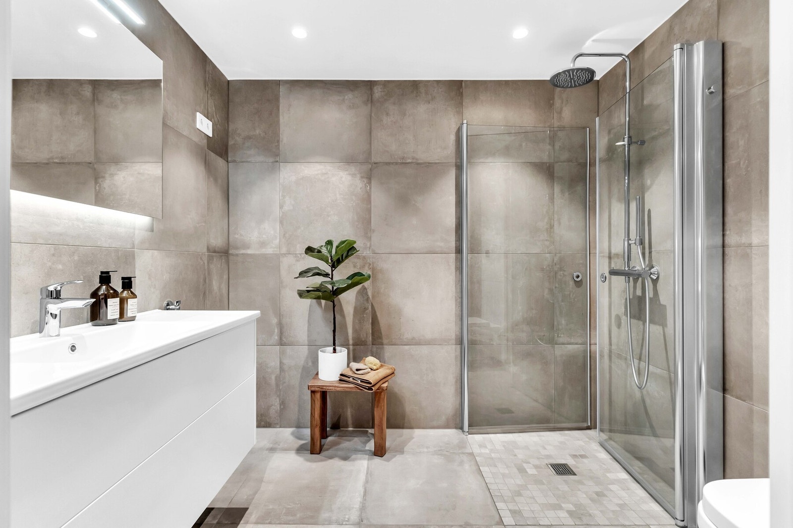 Det største baderommet fremstår med et helhetlig og stilrent design. Store, moderne fliser i nøytrale toner som skaper et tidløst uttrykk Galleribilde