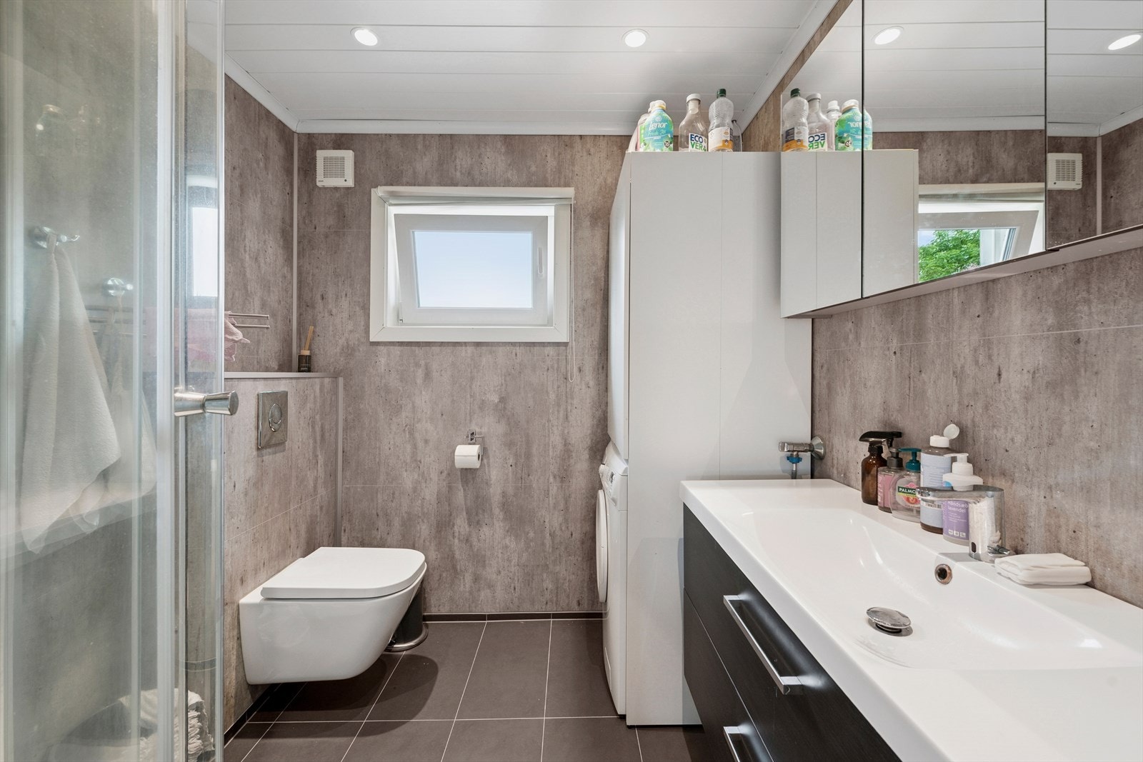 Badet består av speil, servant med seksjon, veggmontert toalett, dusjkabinett og opplegg for vaskemaskin. Galleribilde