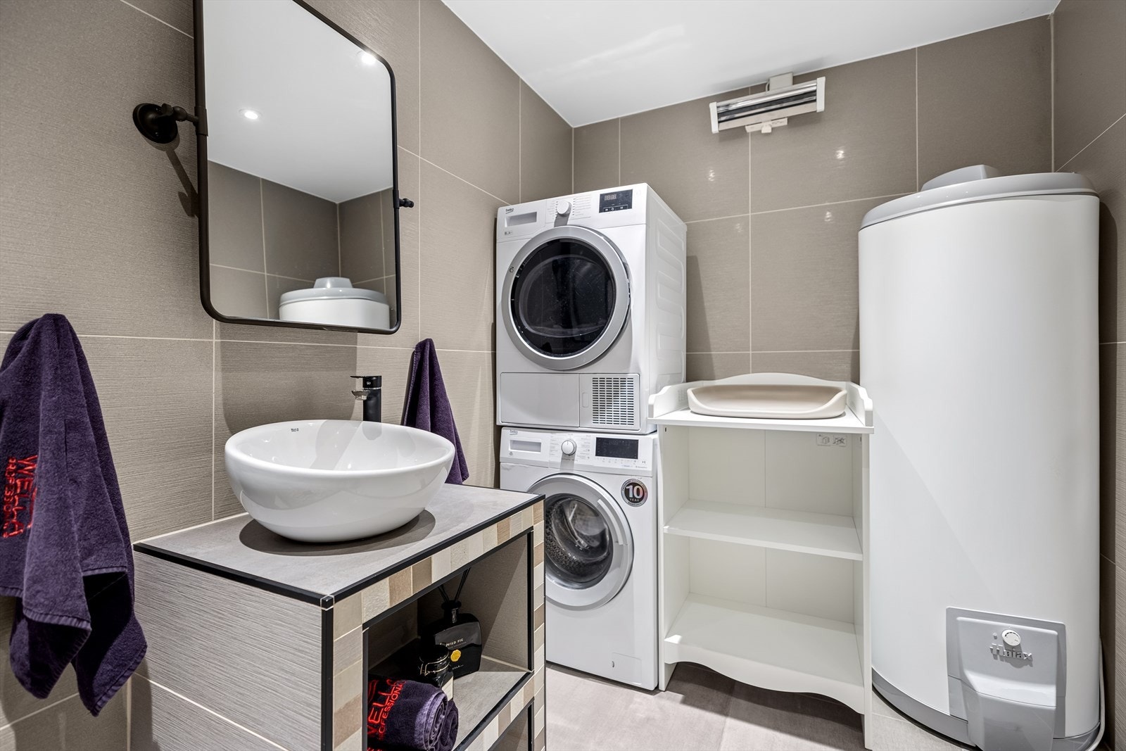 Badet er utstyrt med et gulvstående toalett, en dusjhjørne med glass og svingbare dører, samt en servant på benkeplate med skap under og ettgrepskran. Galleribilde