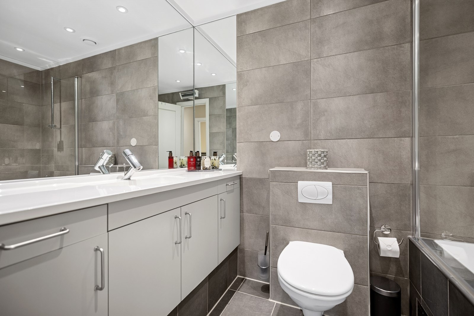 Det andre badet i første etasje er innredet med heldekkende servant med skap og skuffer under, vegghengt toalett med skjult sisterne, et innmurt badekar og en svingbar dusjvegg plassert på badekarskanten. Galleribilde