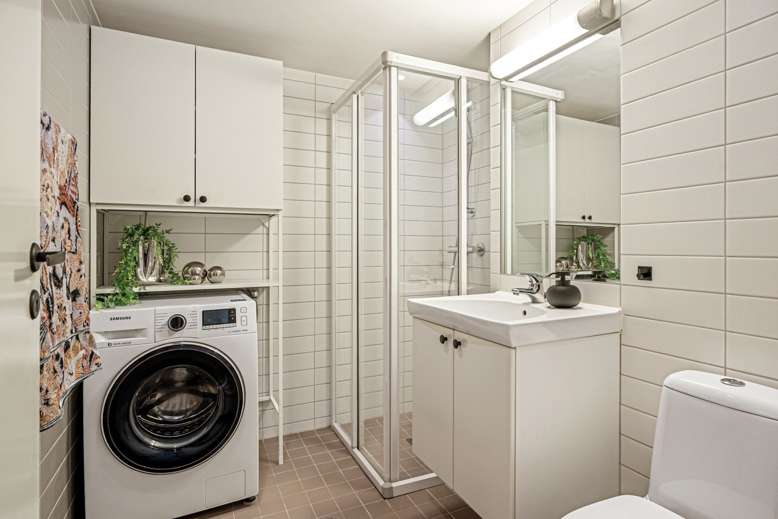 Lyst flislagt bad med varmekabler, toalett, dusjhjørne med skyvedører og opplegg for vaskemaskinRommet har integrert speil med overlys og hvit servant Galleribilde