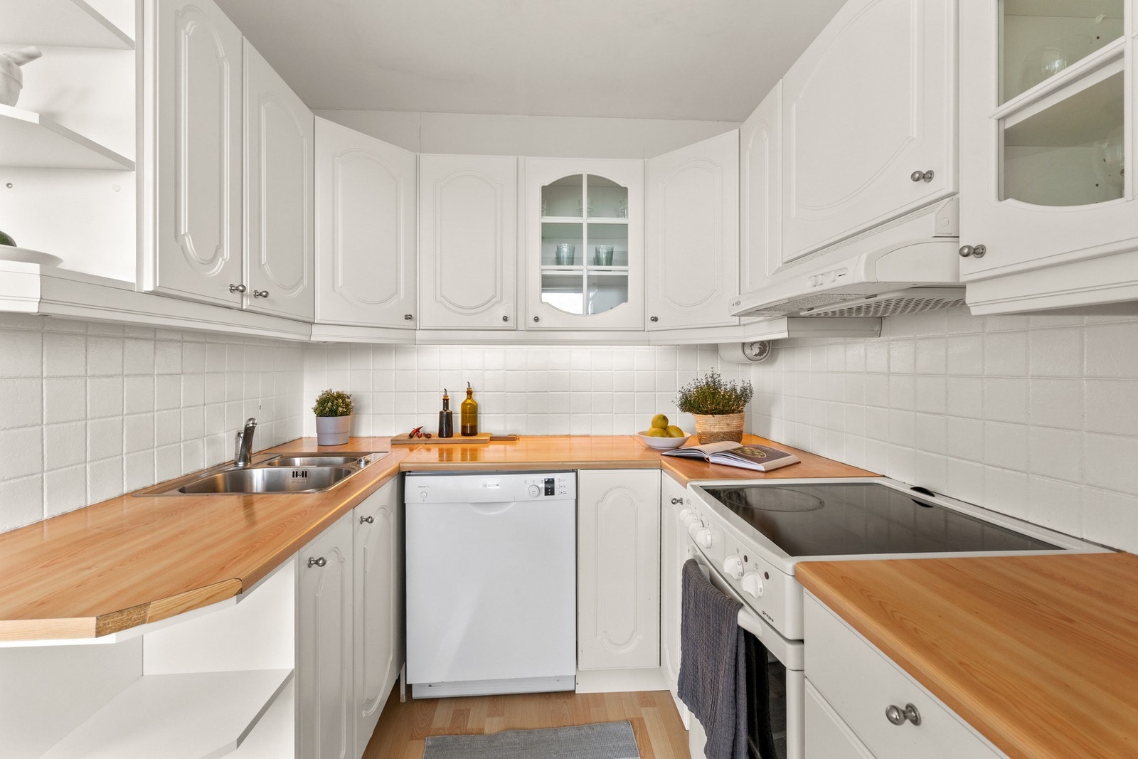 . Videre er kjøkkenet utsyr med nedfelt kum i rustfritt stål, fliser på vegg over benkeplaten og belysning under overskap Galleribilde