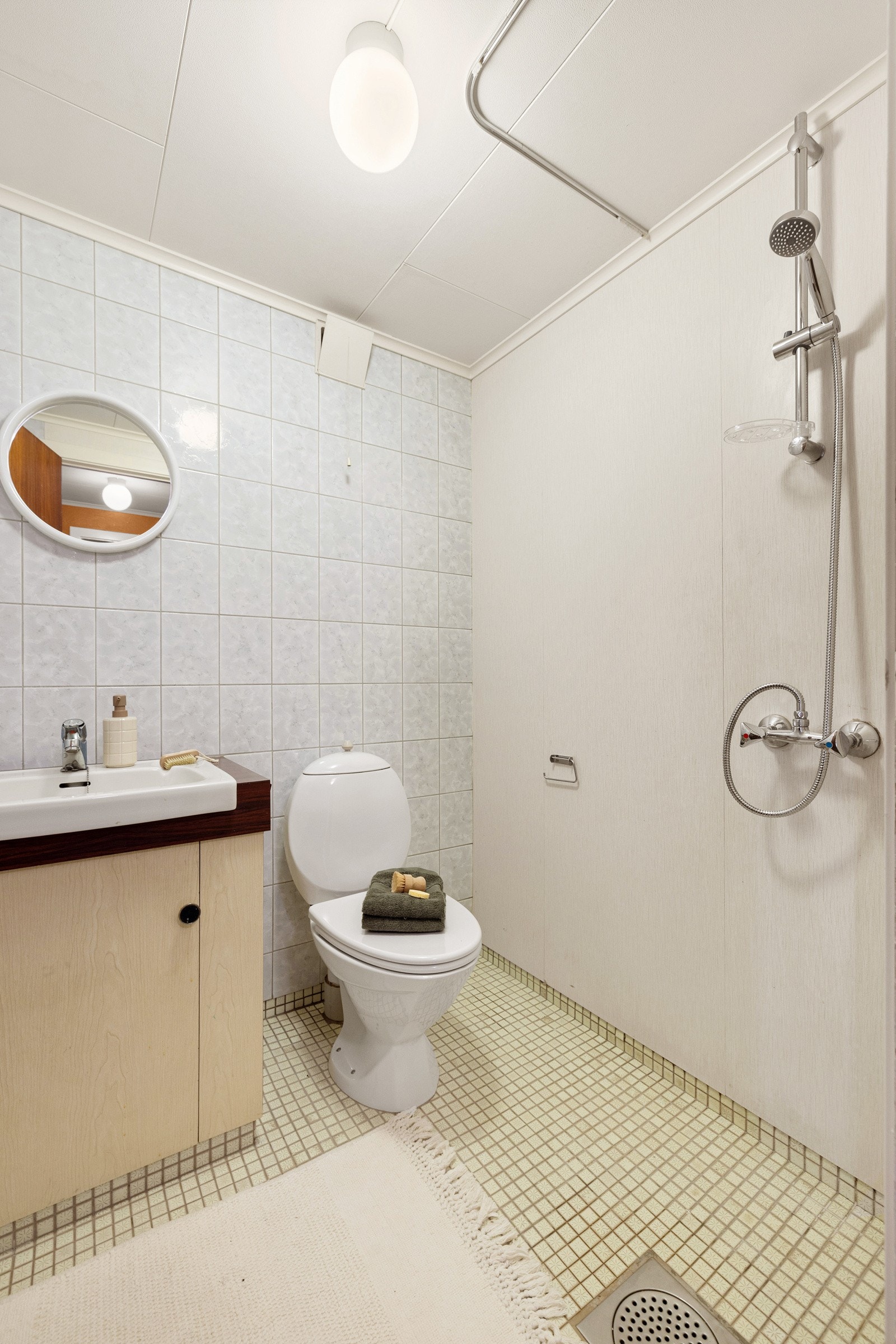 Badet er innredet med gulvstående toalett, servant med underskap og et åpent dusjhjørne uten vegger. Galleribilde