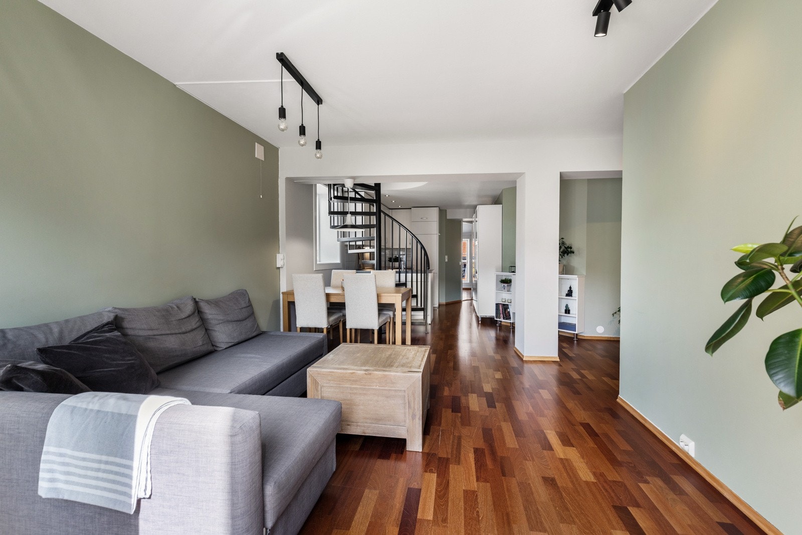 Parkett på gulvet kombineres med vegger malt i lysegrått og en dus grønnfarge. En spiraltrapp med flislagte trinn fungerer som romdeler mellom stuen og kjøkkenet. Galleribilde