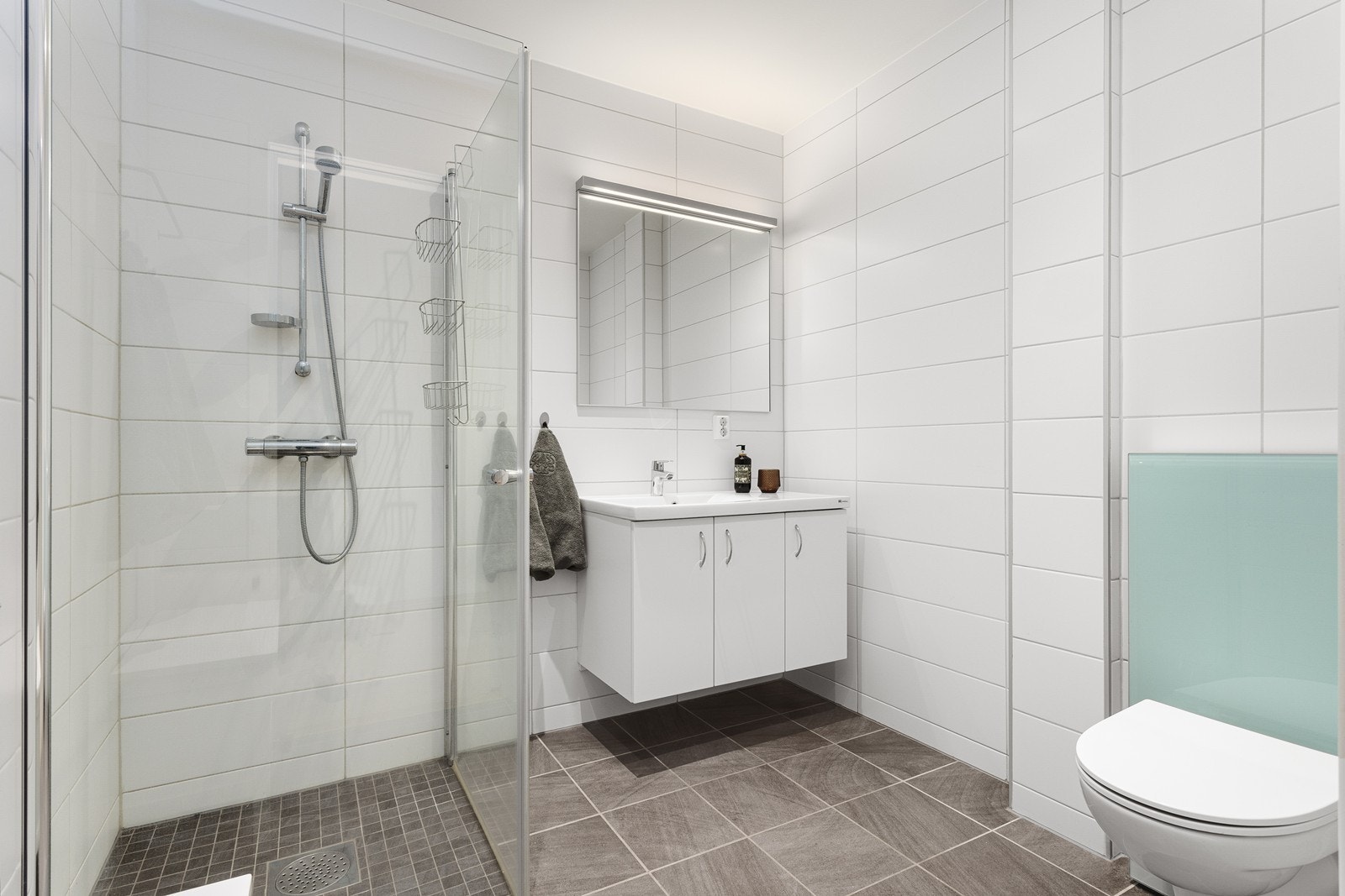 Stilrent og moderne flislagt bad med varmekabler i gulv. Baderomsinnredningen er lys med heldekkende nedfelt servant og rom for oppbevaring i underskap og speil med belysning. Videre har badet dusjhjørne med dus Galleribilde